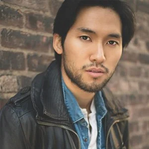 A man with dark hair and a beard wearing a leather jacket and denim shirt standing against a brick wall.