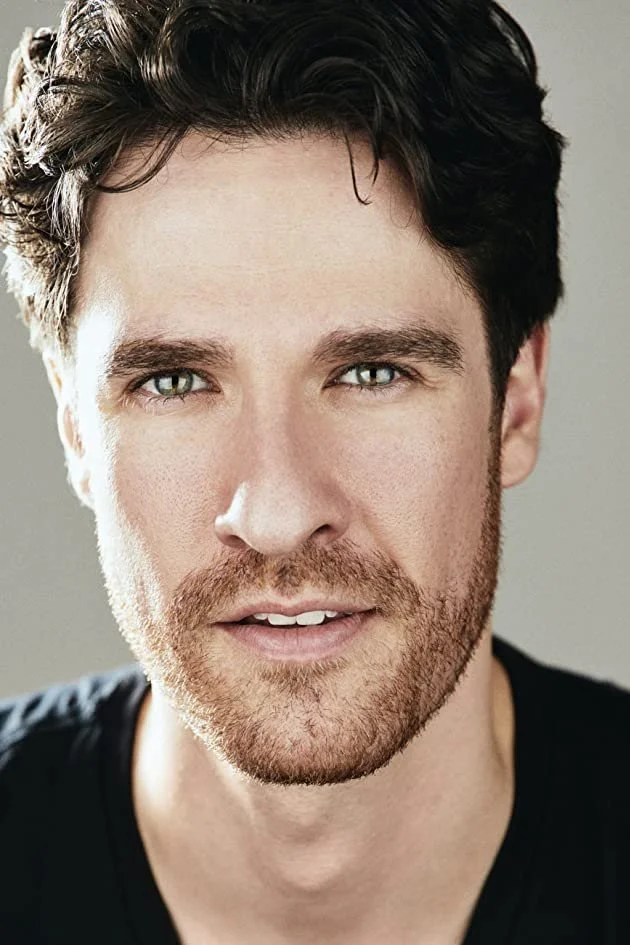 Close-up of a man's face with brown hair and beard, wearing a black shirt, looking directly at the camera.
