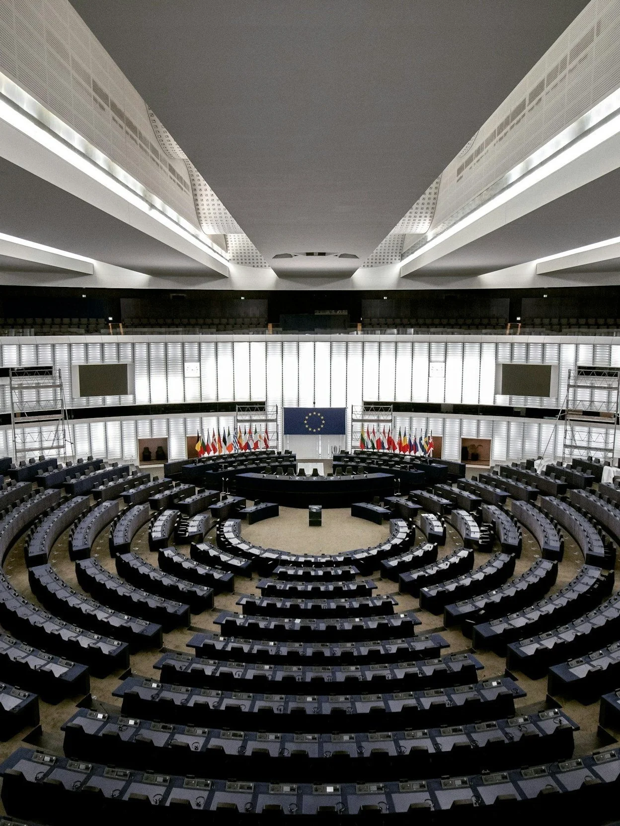 Interior of a large circular parliamentary chamber with rows of seating, desks, a central podium, and flags on display.