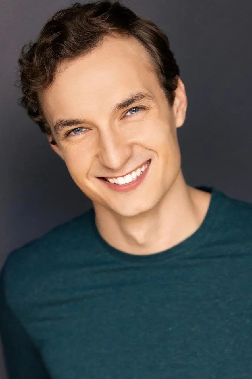 Smiling person with short curly hair and a green shirt against a dark background.