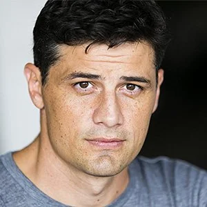 Portrait of a man with dark hair looking directly at the camera, wearing a gray shirt against a neutral background.