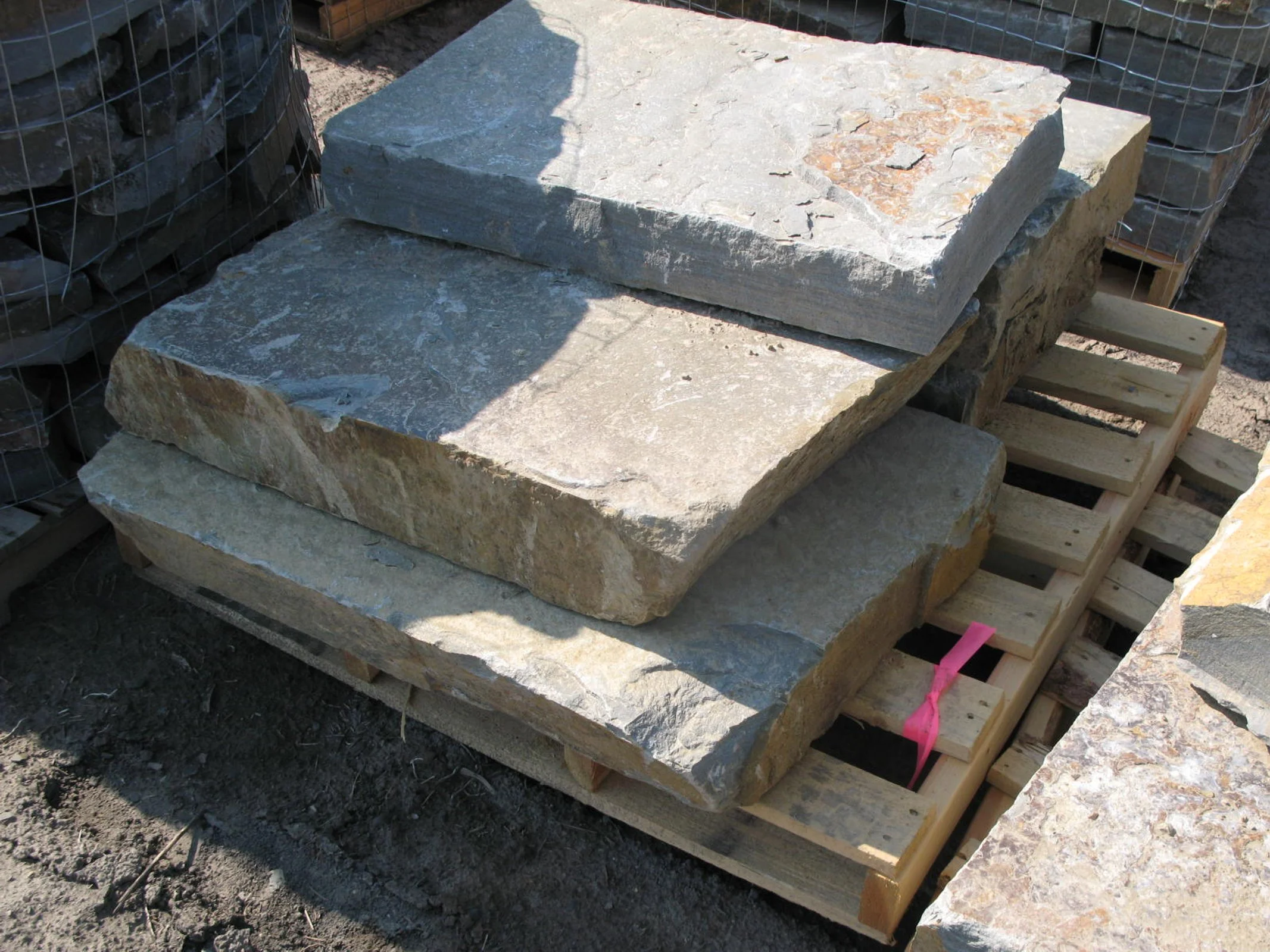 Large natural stone slabs stacked on a wooden pallet outdoors, with soil and fencing in the background.