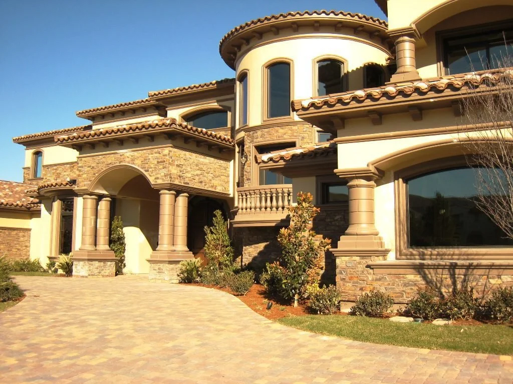 Luxury house with stone and stucco exterior, arched doorway, columns, bay windows, and a tiled roof, set on a paved driveway with landscaping.