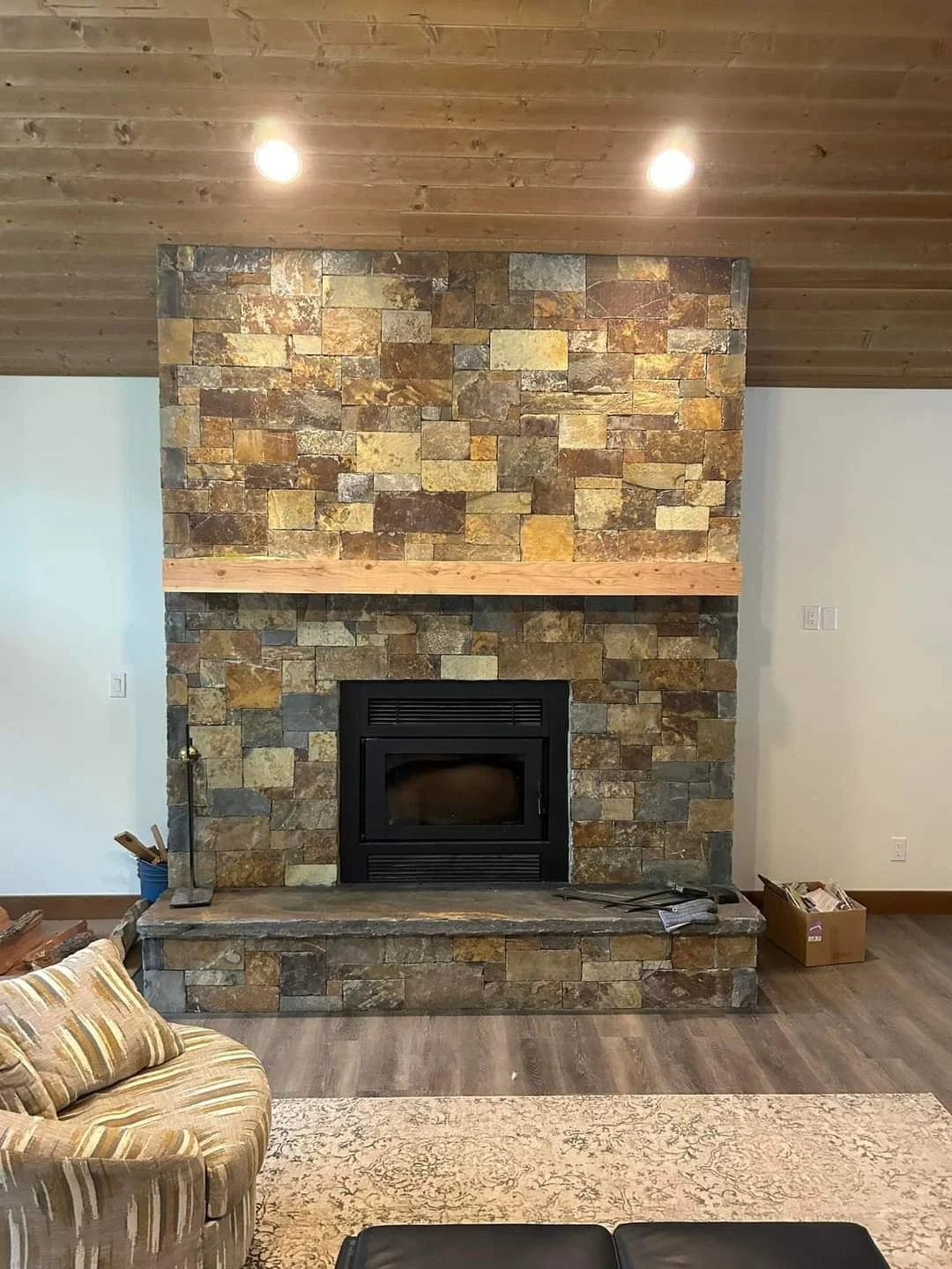 Living room with a stone fireplace, wooden mantle, hardwood floor, beige patterned rug, and partially visible furniture.