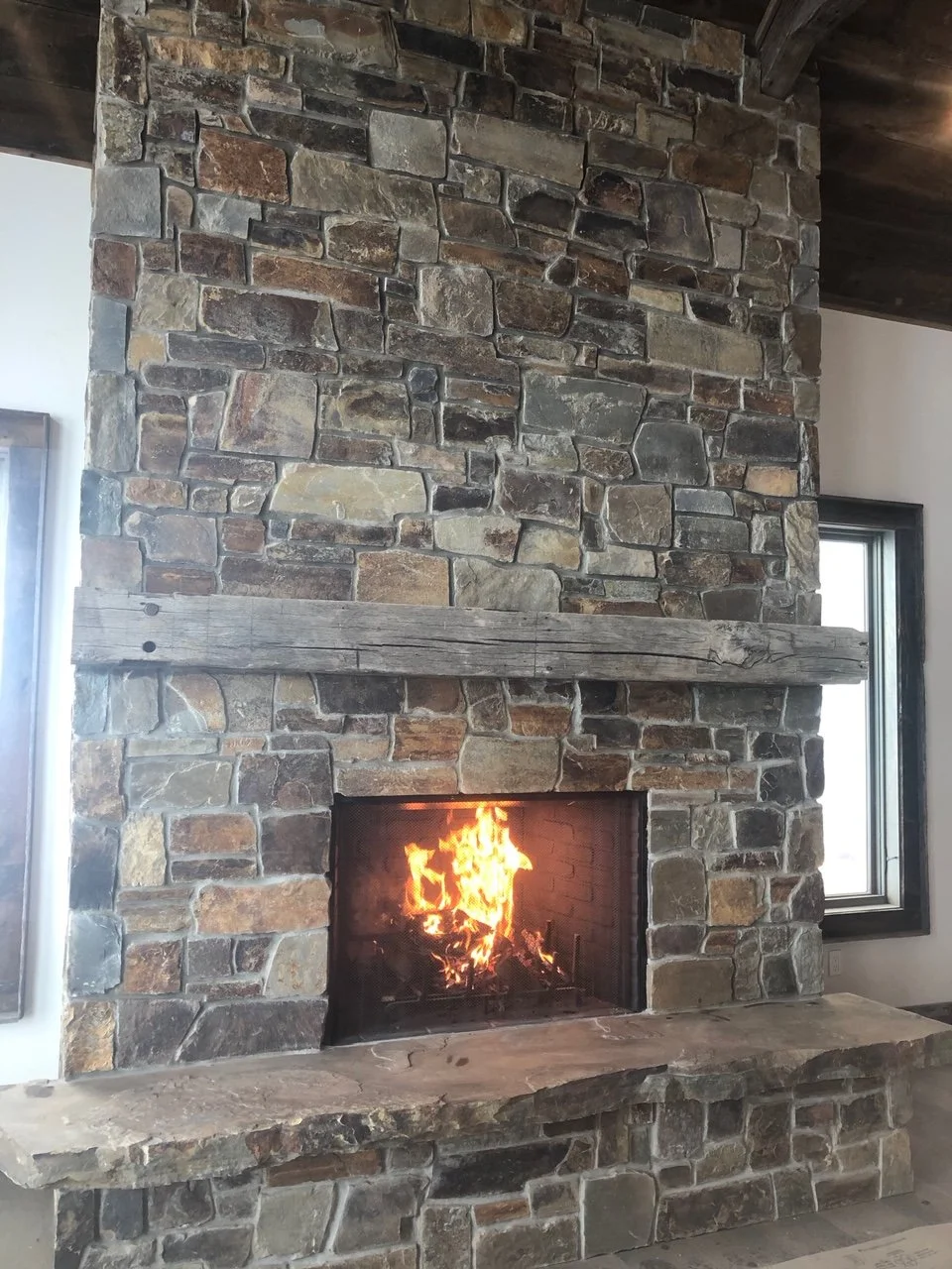 A large stone fireplace with a burning fire, a wooden mantel, and side windows in a cozy living room.
