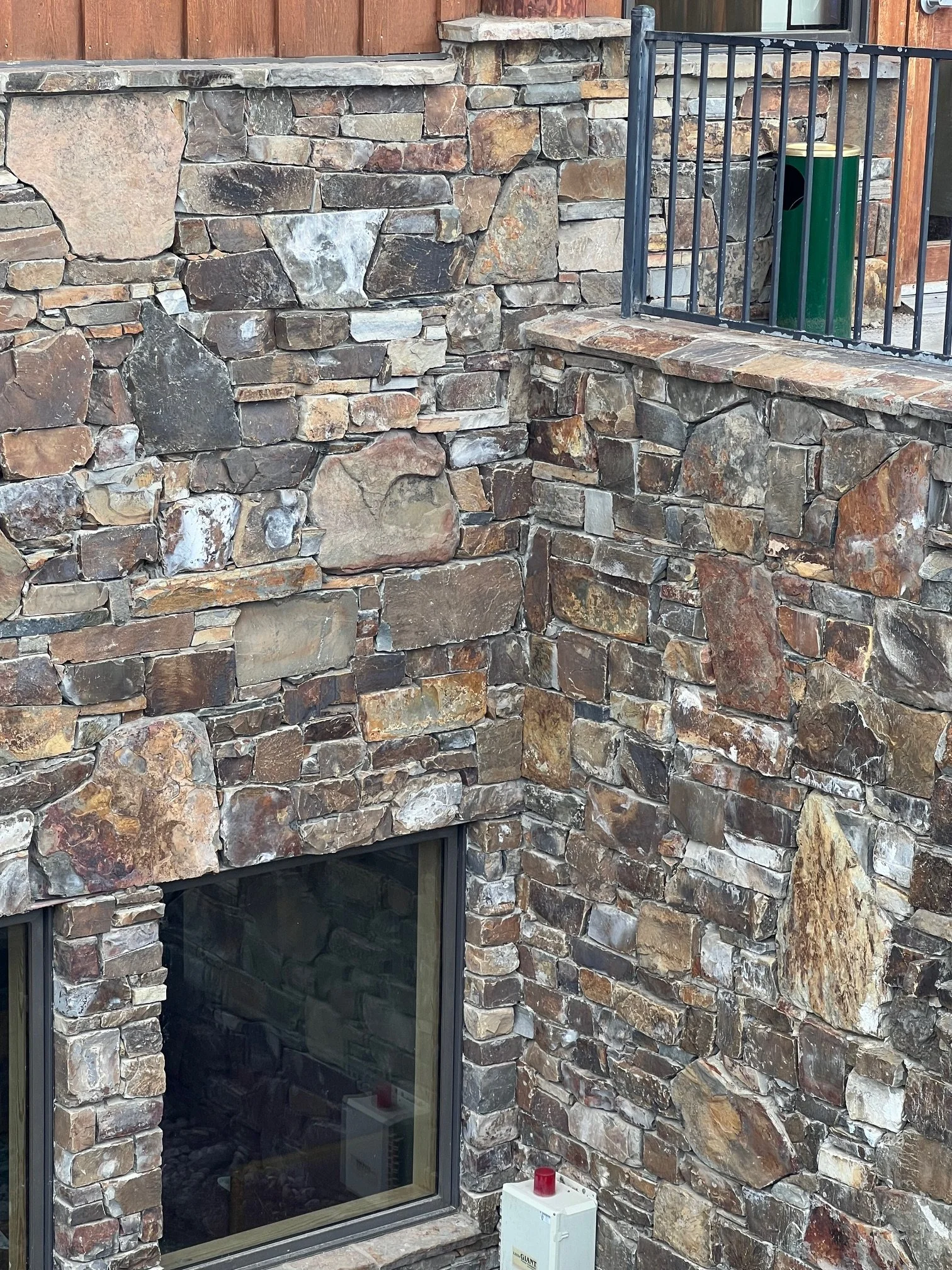 A stone exterior wall of a building with a large window. The wall is made of irregularly shaped multicolored stones. There is a black metal railing by the window and a green recycling bin behind it. A white box with a red light on top is on the groun