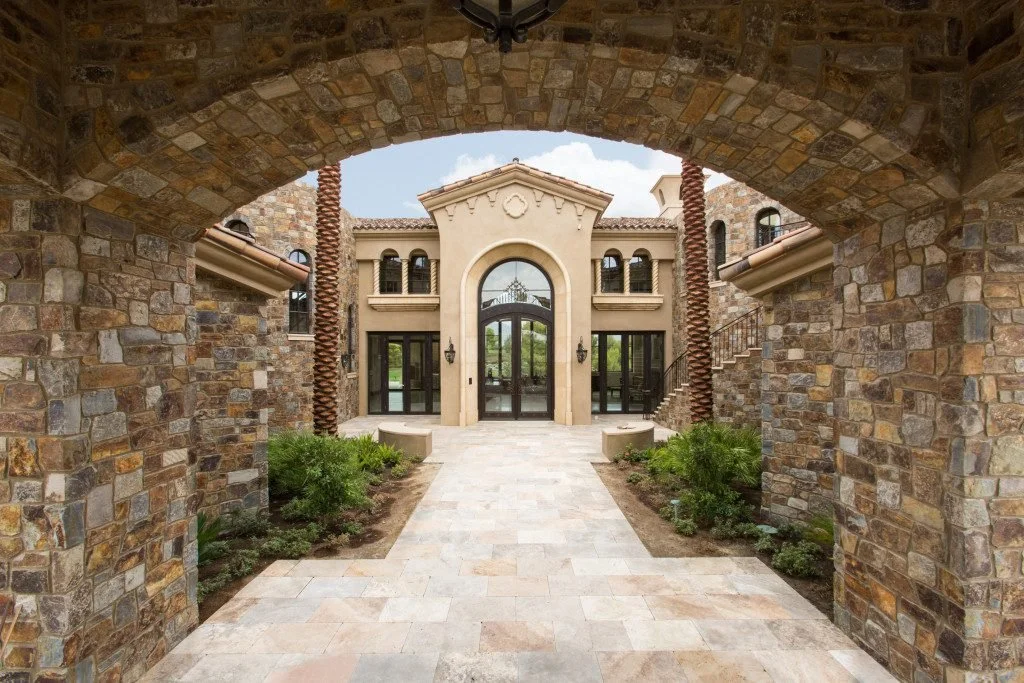Front entrance of a luxurious house with stone archway, paved walkway, and elegant building facade with large glass doors and windows.