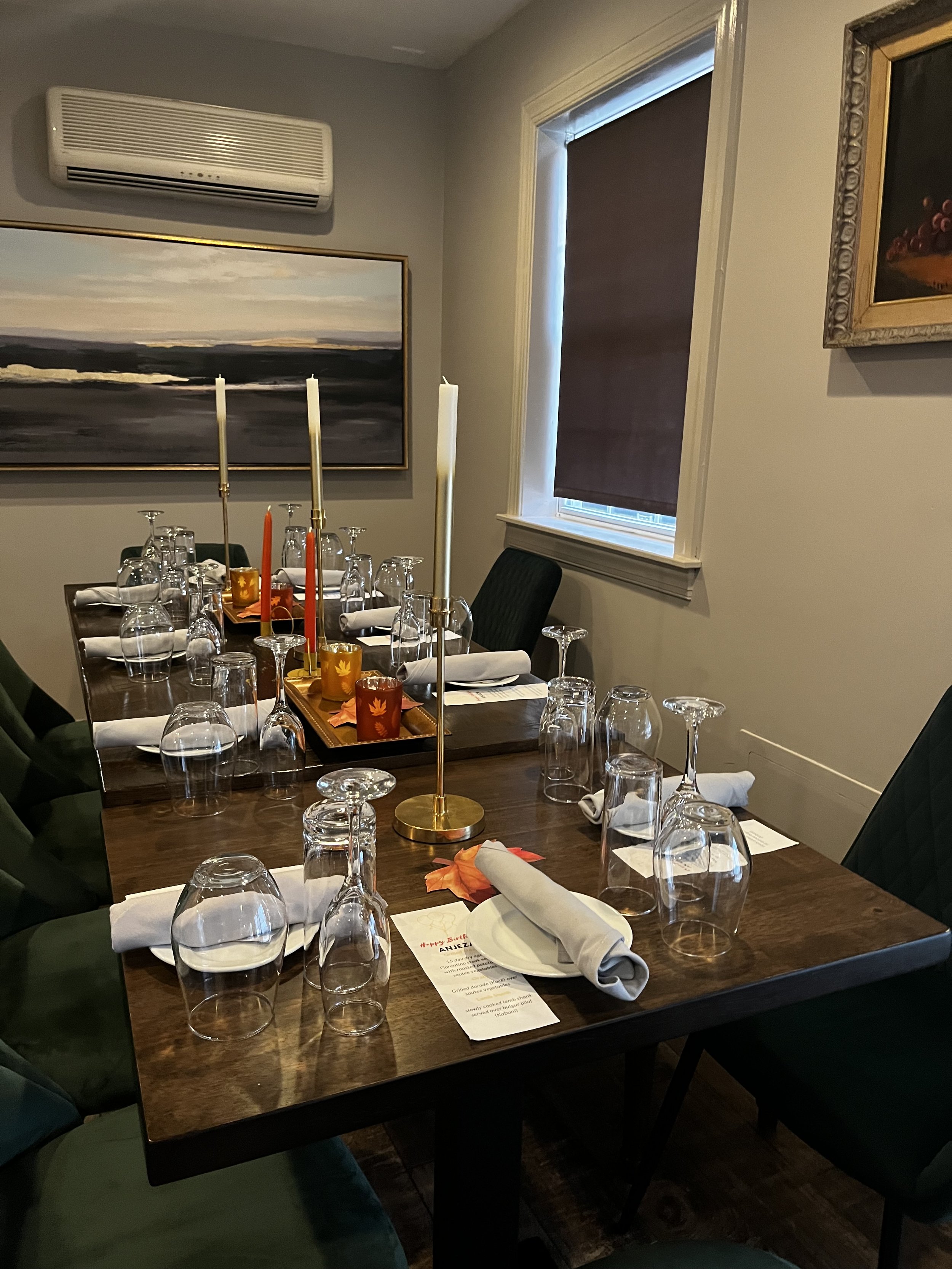 A dining table set for a meal with wine glasses, cloth napkins, candles, and a menu, in a room with framed paintings and an air conditioning unit.
