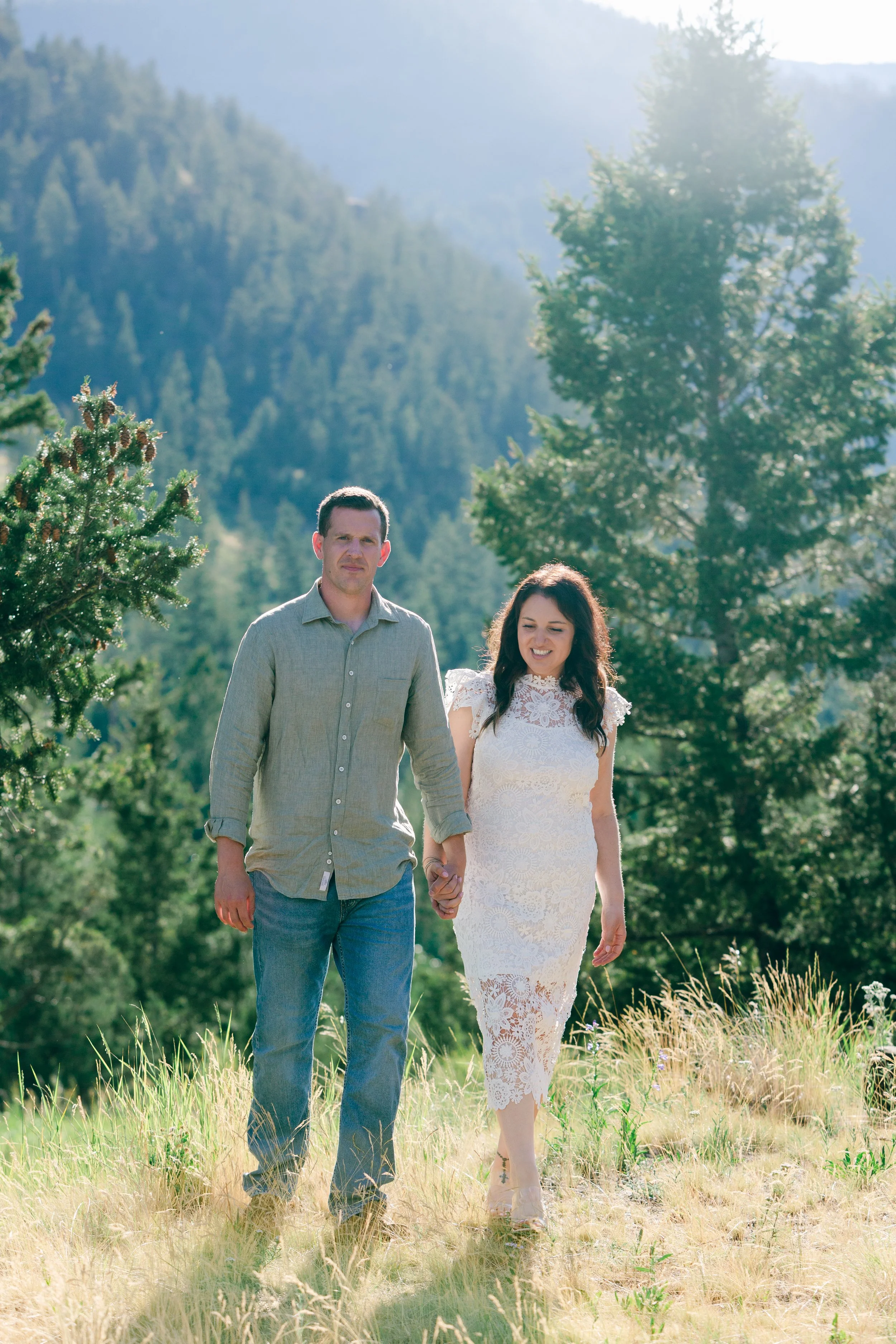 Couple holding hands and walking through a meadow during an engagement session at Wayfarers Chapel near Cody, Wyoming, with the Absaroka Mountains in the background.