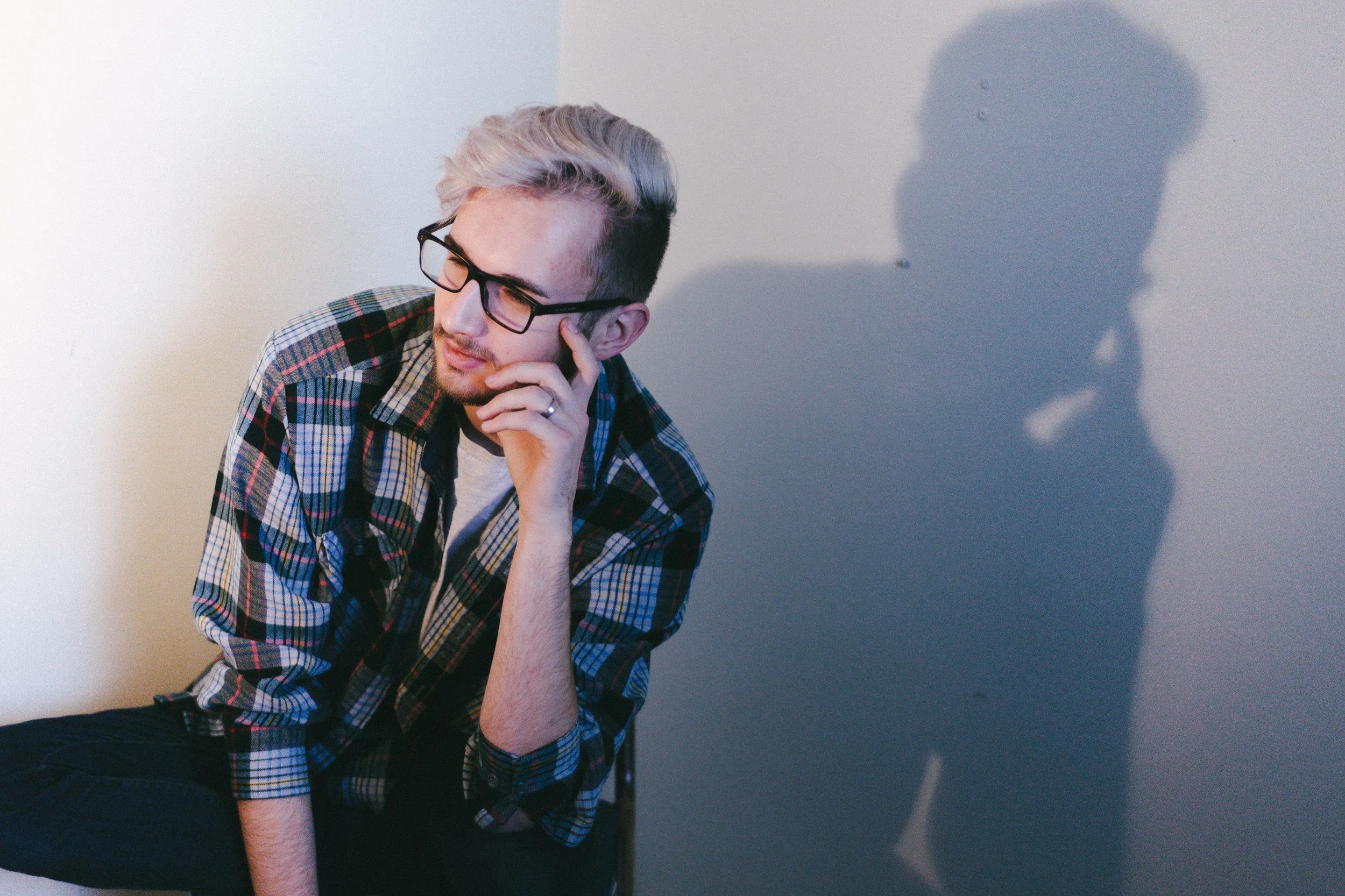 A young man with glasses and blond hair with dark roots, wearing a plaid shirt, sitting with his hand resting on his face, looking contemplative. His shadow appears on the wall behind him.