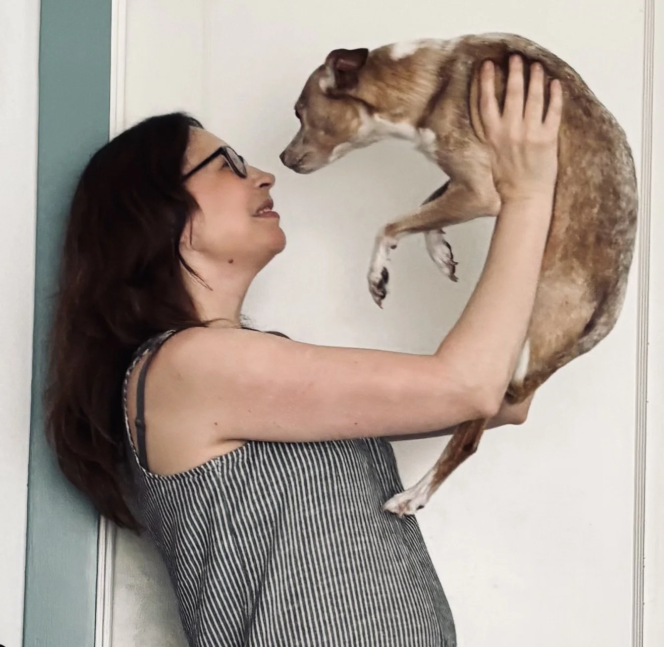 A woman with long dark hair and glasses holding a small brown and white dog in front of a light-colored wall.
