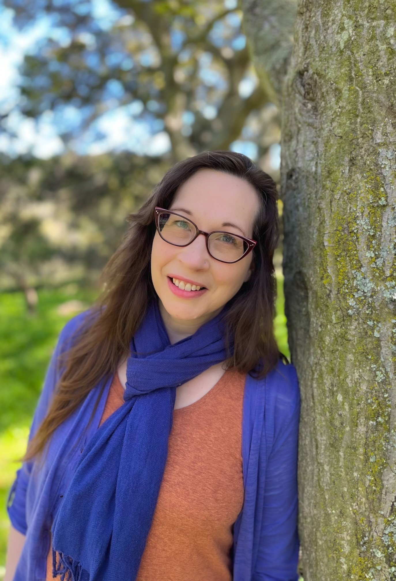 A woman with glasses and a nose piercing smiling outdoors next to a tree, wearing a blue scarf and an orange shirt with a blue jacket, in a park setting.