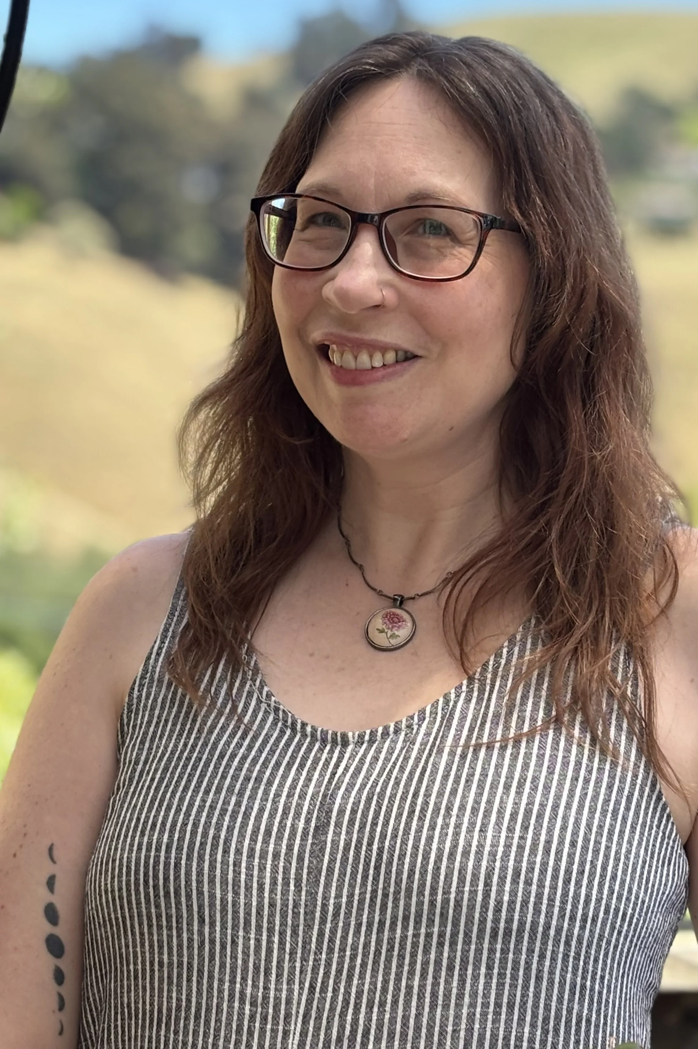 A woman with glasses and a necklace smiling outdoors with a blurred background of trees and sky.