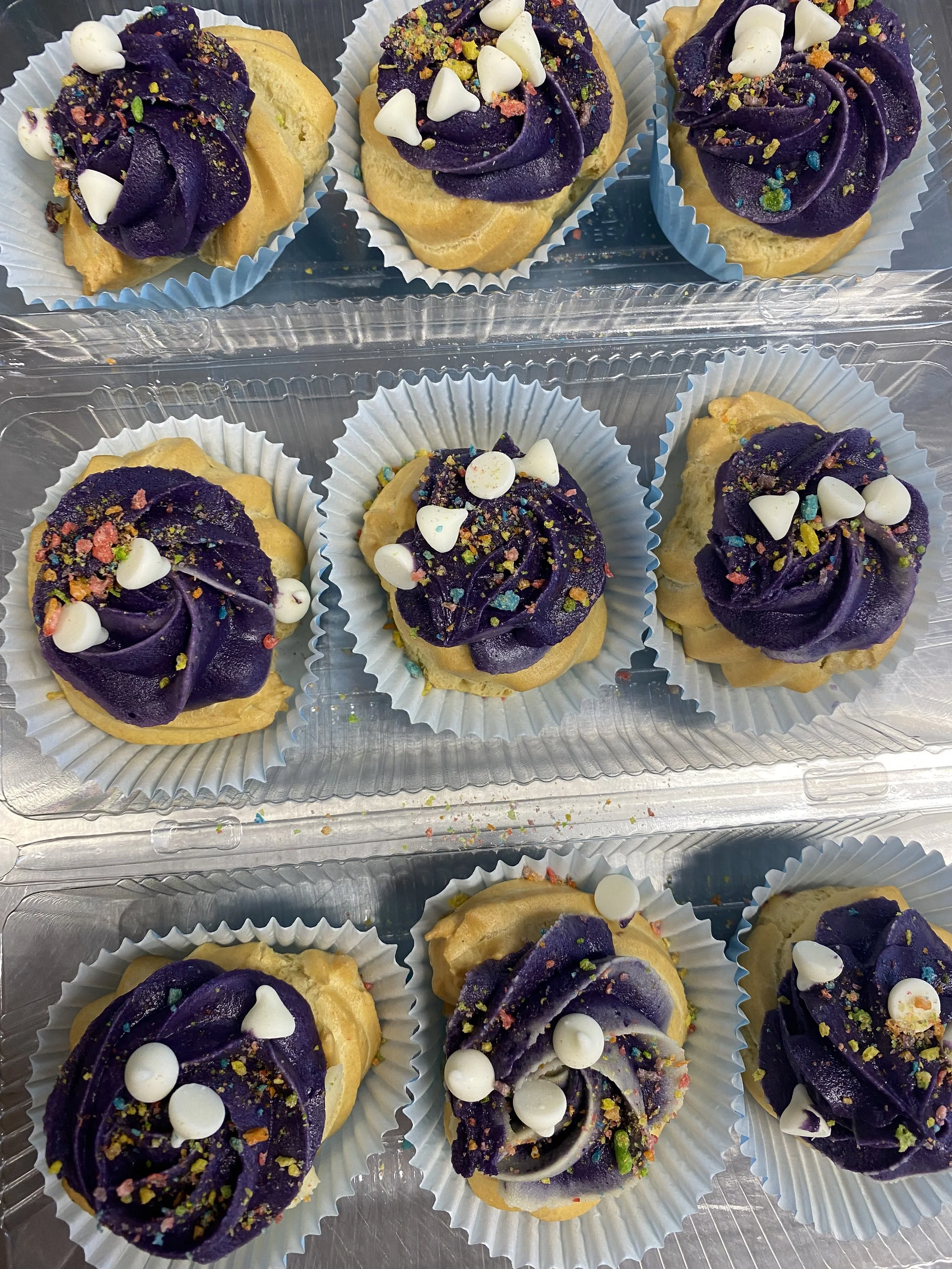 Nine cupcakes with purple frosting and white heart-shaped and round sprinkles in paper cups, arranged in three rows inside a plastic container.