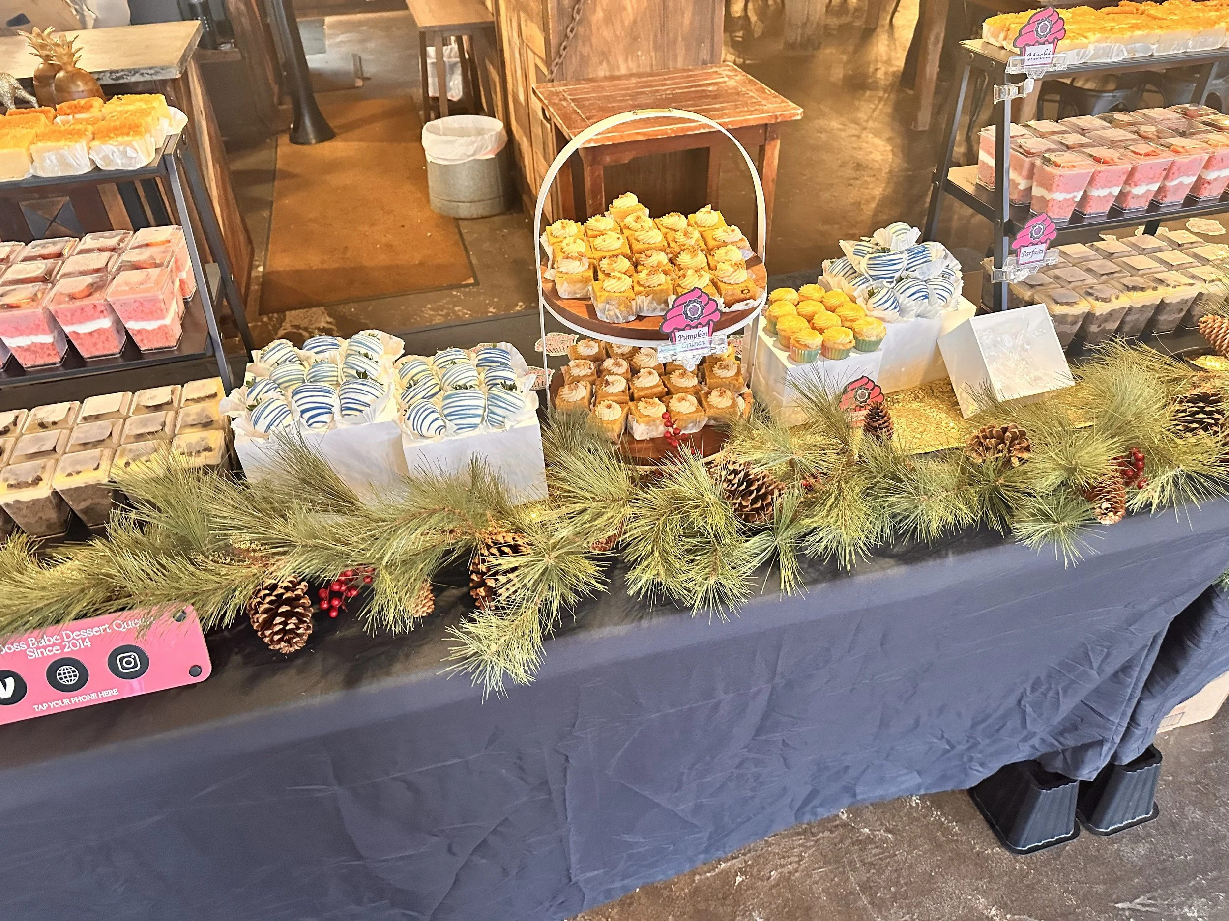 Dessert table decorated with pine branches and pine cones, featuring various cupcakes and small dessert cups.