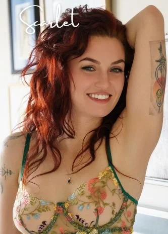 Close-up of a woman with red hair lying on a bed, looking at the camera. She has tattoos on her arm and wears black lingerie. The word 'Scarlet' is written in white cursive text in the top left corner.