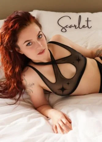 Close-up of a woman with red hair lying on a bed, looking at the camera. She has tattoos on her arm and wears black lingerie. The word 'Scarlet' is written in white cursive text in the top left corner.