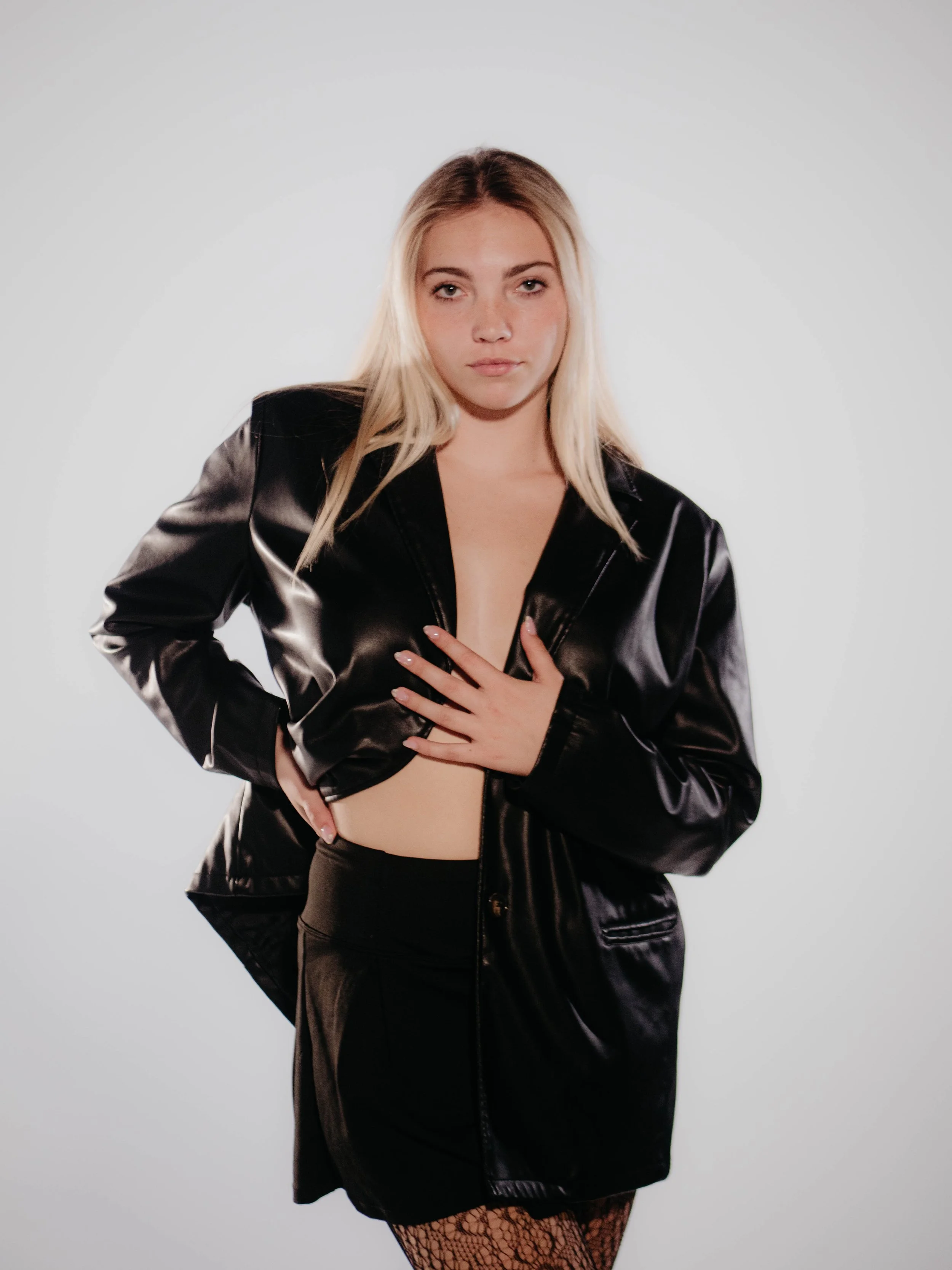 A young woman with blonde hair and fair skin, wearing a black satin jacket, black skirt, and fishnet stockings, standing against a plain white background.