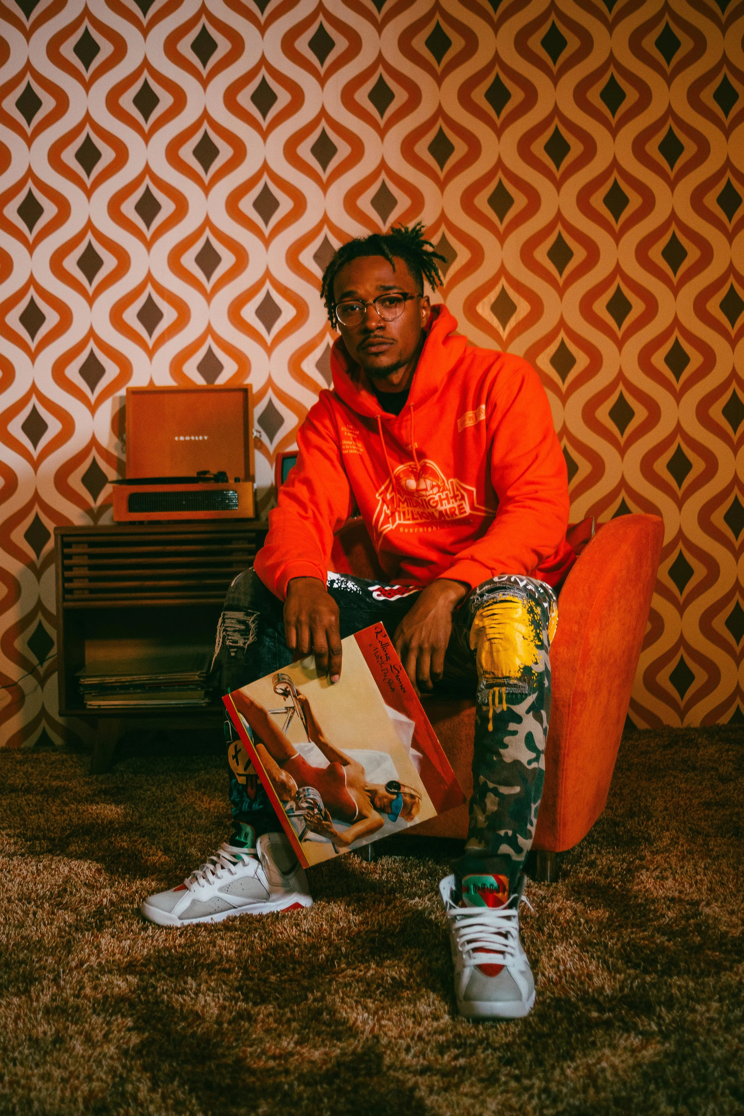 A man sitting in an orange armchair holding a vinyl record, with a vintage turntable and record shelf in the background, against a retro patterned wallpaper.