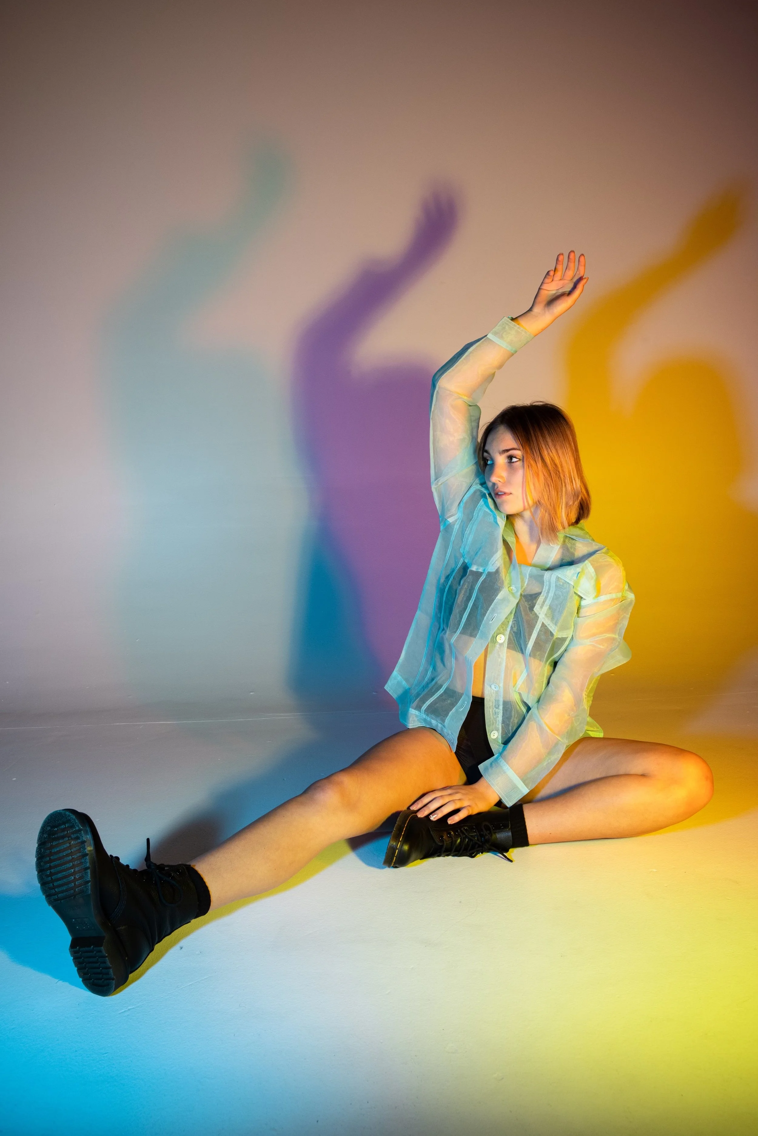 Young woman sitting on the floor in a colorful lighting setting, wearing a transparent blouse and black boots, with her arm raised and her shadow cast on the wall behind her.