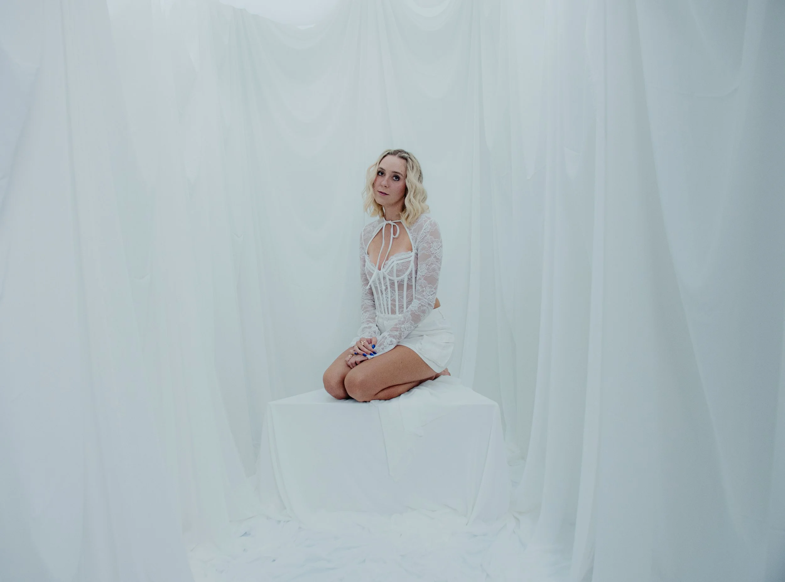 A woman in white lace clothing sitting on a white block in a room with white draped fabric walls.