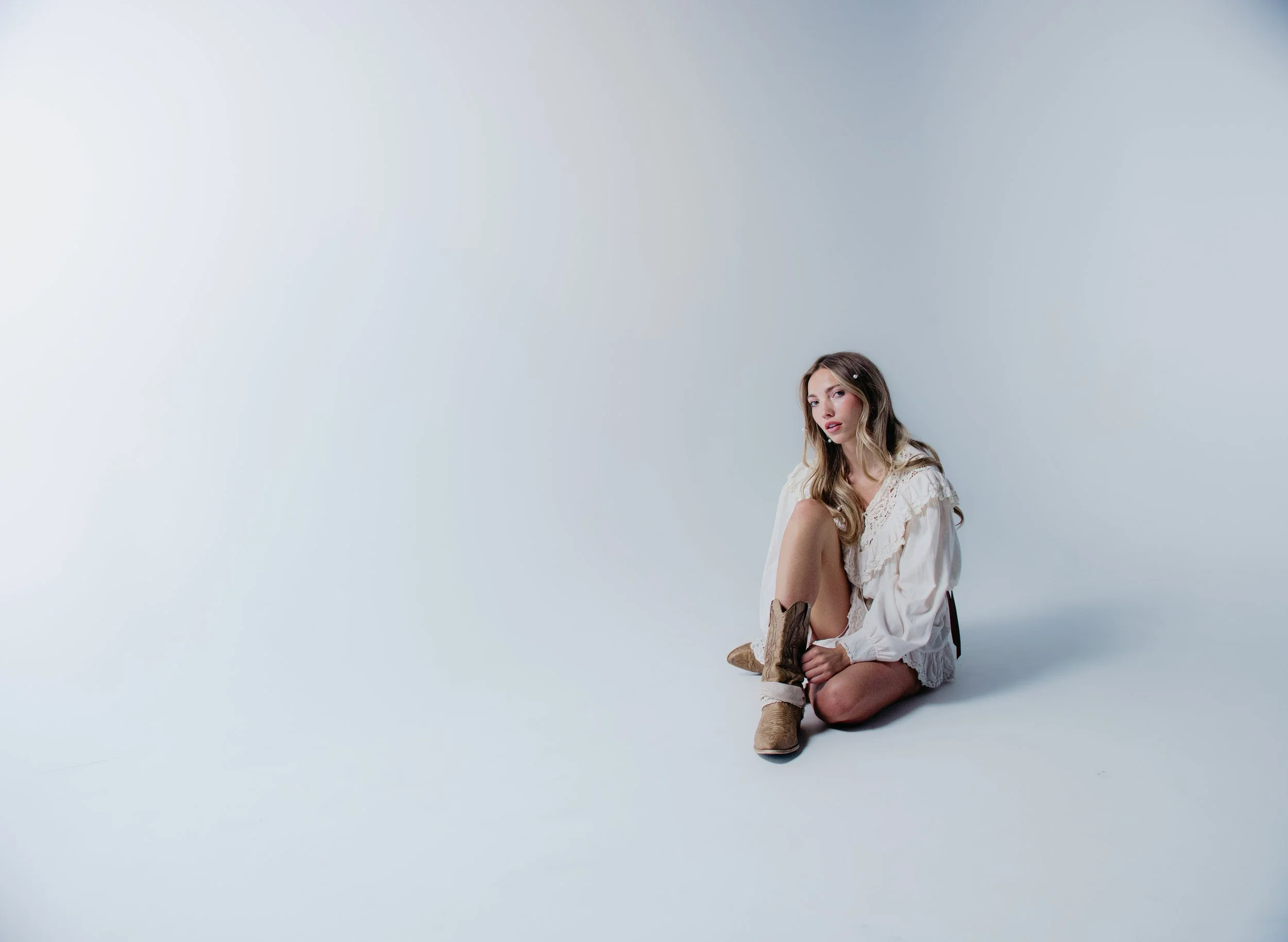 A woman with long brunette hair, wearing a white dress and cowboy boots, sitting on the floor against a plain light background, looking at the camera.