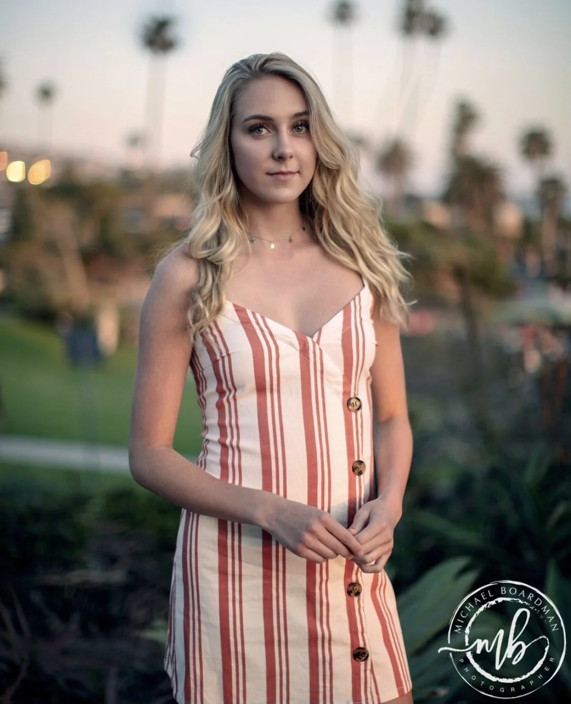 A young woman with long blonde hair in a striped sleeveless dress stands outdoors at sunset, with palm trees and a cityscape in the background.