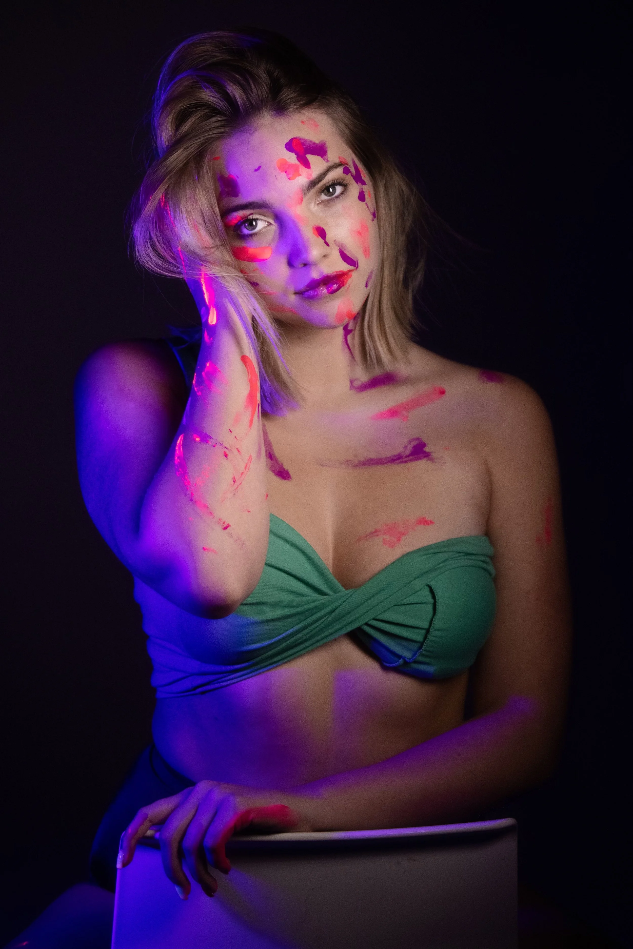 A woman with short blonde hair posing against a black background, illuminated with purple and pink light, with colorful paint splattered on her face and chest, wearing a green strapless top.