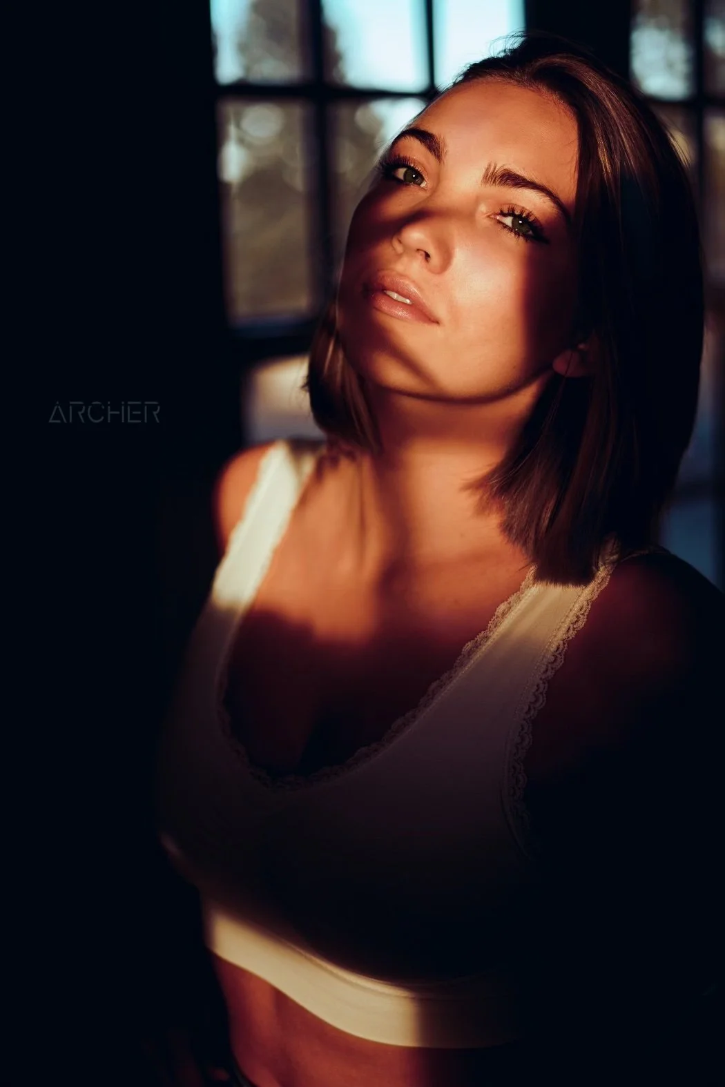 A young woman with short brown hair and green eyes looks at the camera with sunlight casting shadows across her face. She is wearing a white tank top with lace trim and is indoors near a window.