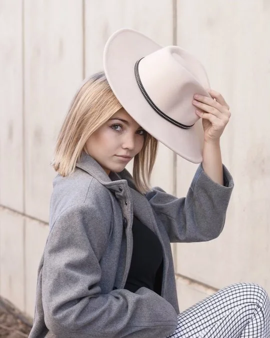A young woman with blonde hair wearing a gray jacket, black top, and checkered pants, holding a white wide-brimmed hat with her right hand, posing outdoors in front of a wooden wall.