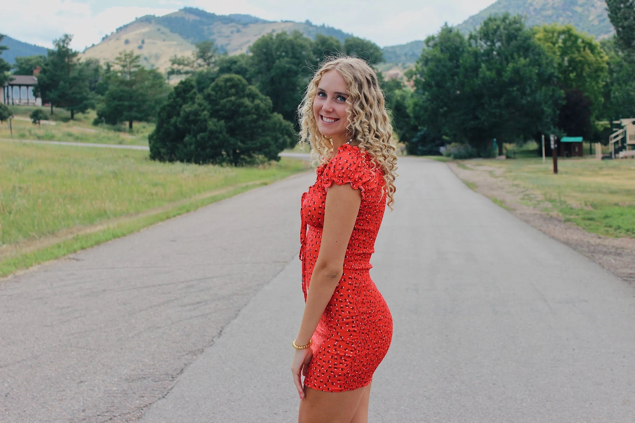 Young woman with curly blonde hair wearing a red dress with small black patterns standing on a paved road in a rural area with green trees and mountains in the background.