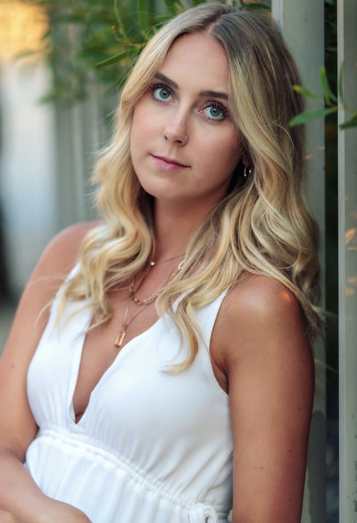 A young woman with blonde, wavy hair and blue eyes posing outdoors near a green fence, wearing a white sleeveless dress and layered gold necklaces.