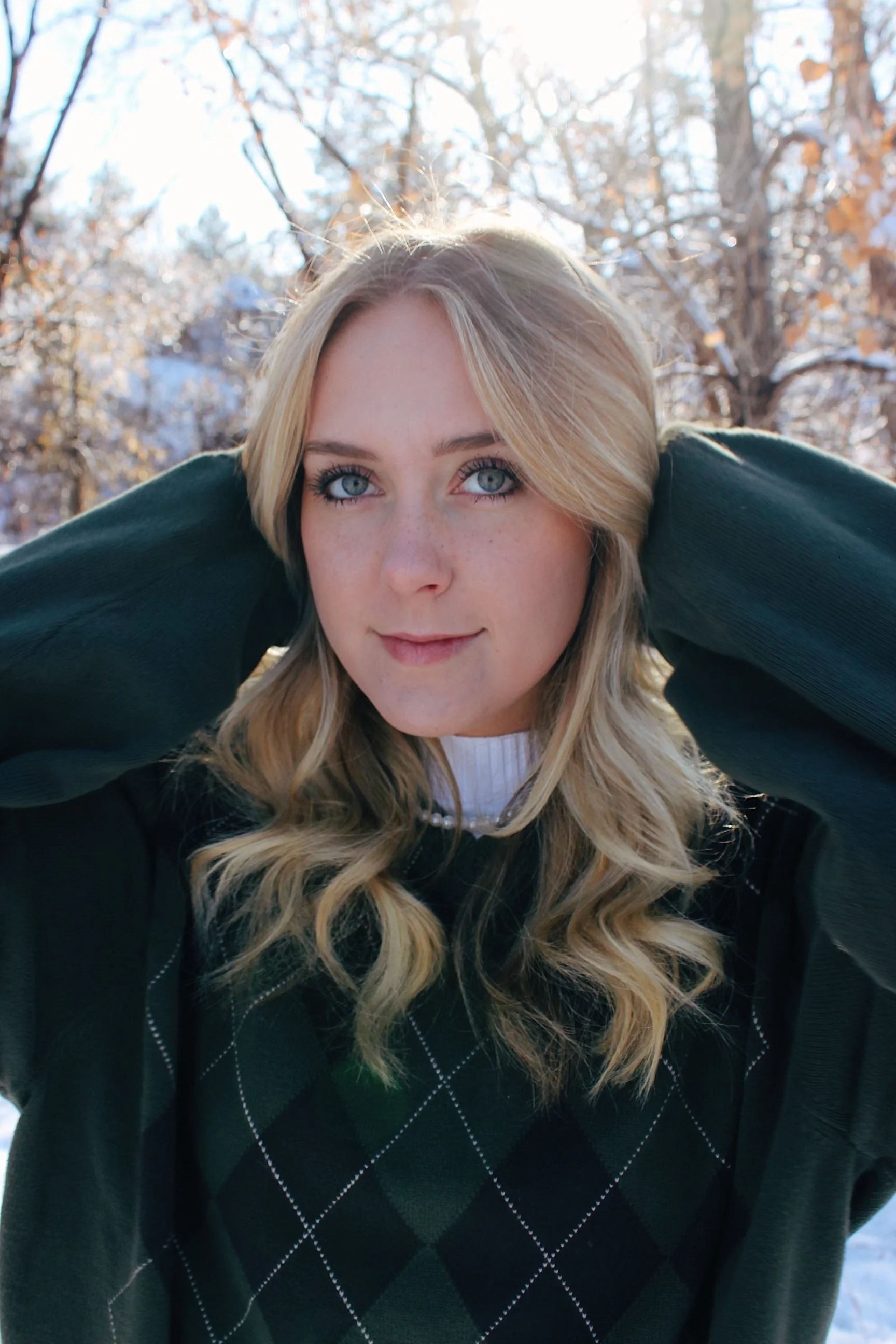 A young woman with blonde hair and blue eyes standing outdoors in a snowy landscape, holding her hands behind her head and looking at the camera.
