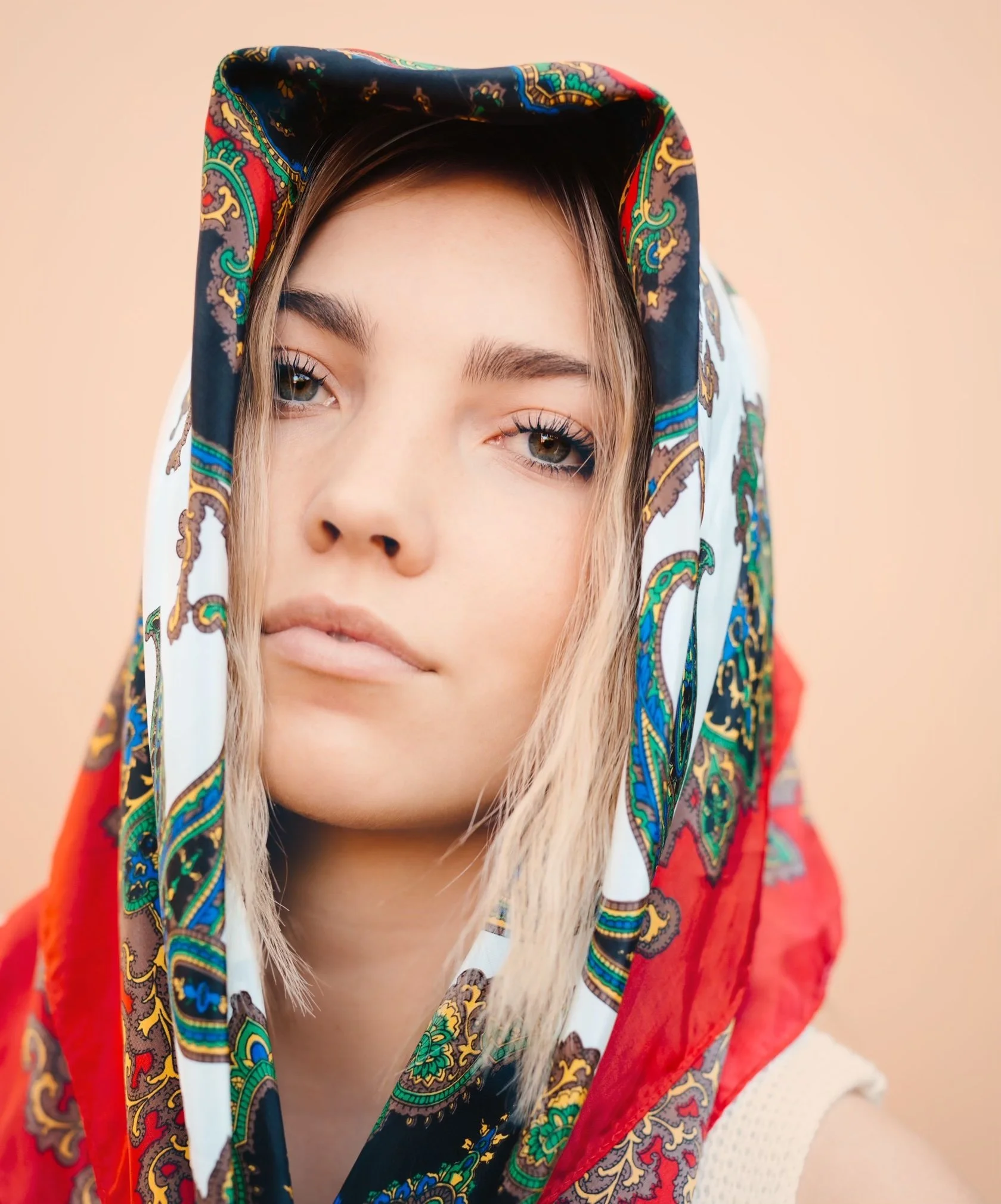 A young woman with blond hair wearing a colorful patterned scarf over her head, with a neutral expression on a peach background.