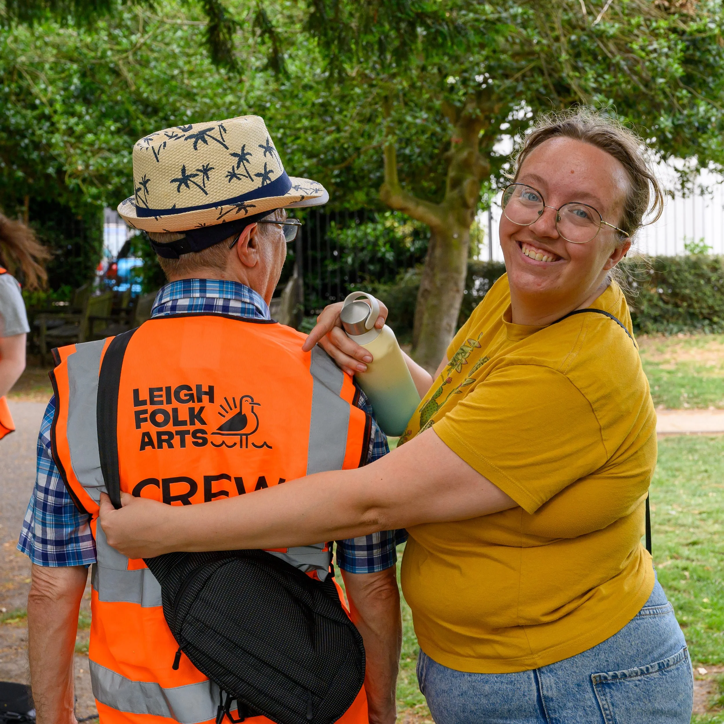 Smiling woman in glasses embraces a volunteer wearing a straw hat and orange Leigh Folk Festival vest outdoors.