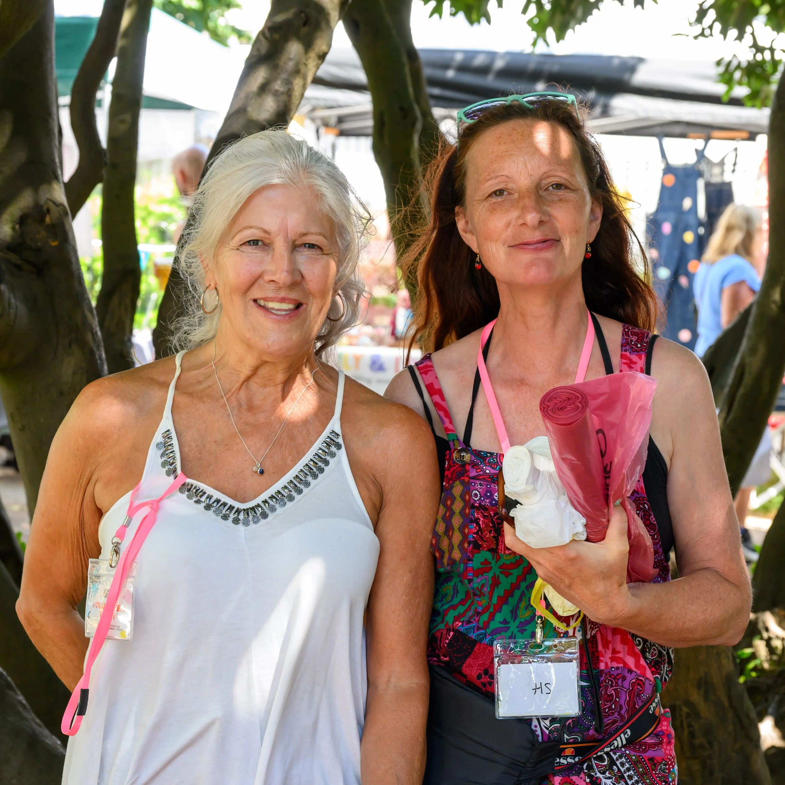 Two smiling women posing outdoors beneath a tree at a colorful event