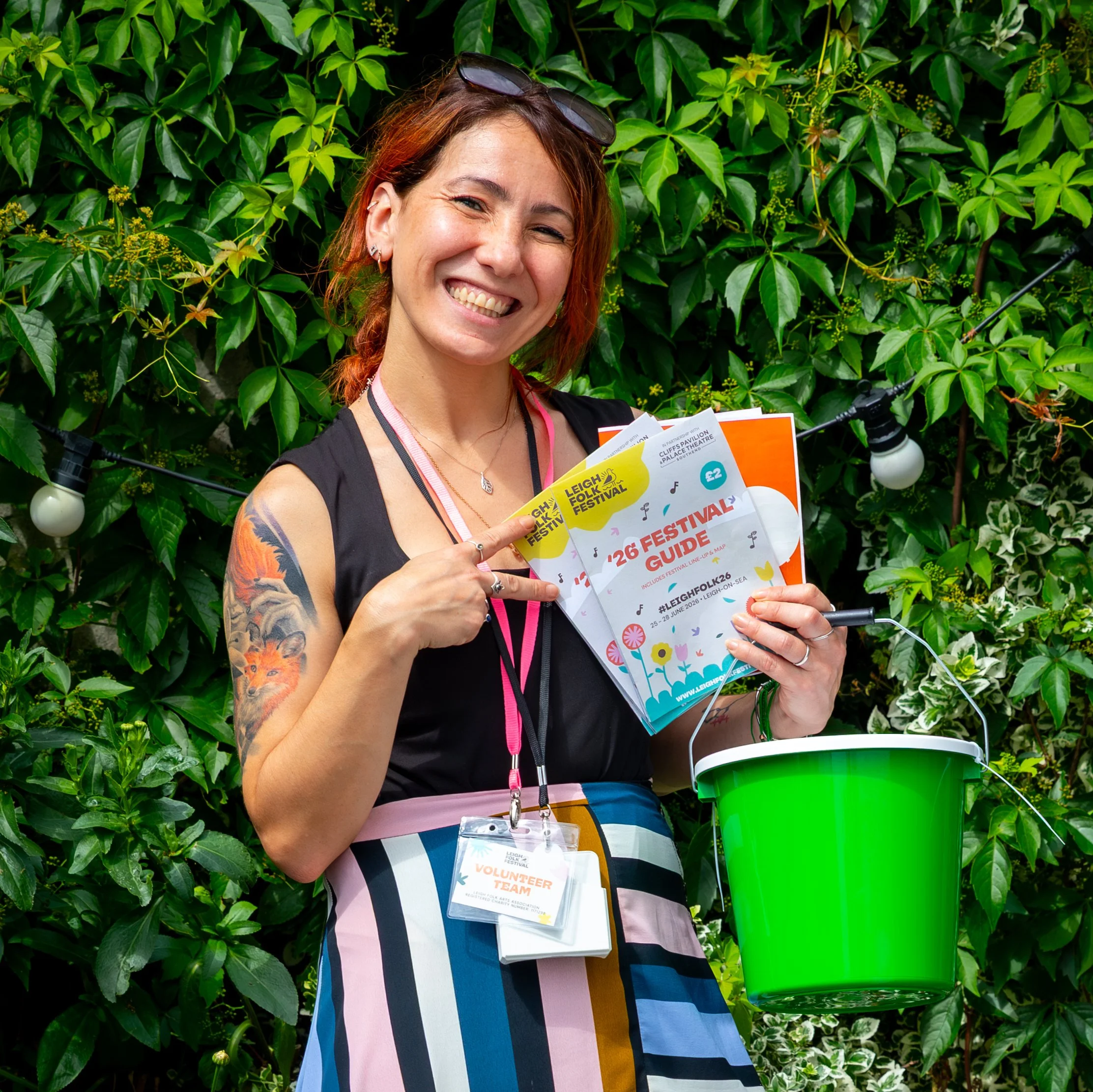 Smiling festival volunteer holding a Leigh Folk Festival guide and green bucket among lush foliage.