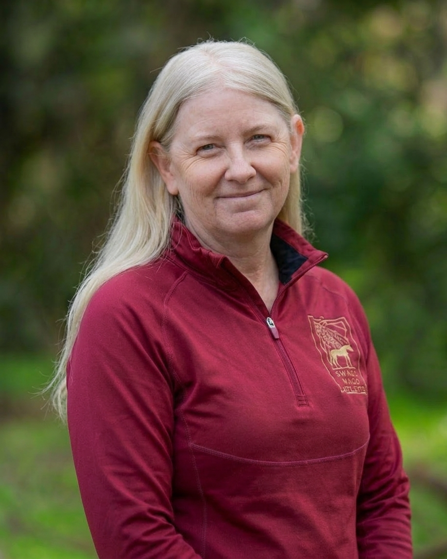 A smiling woman with long, blonde hair wearing a gray sweatshirt with red lettering standing outdoors.