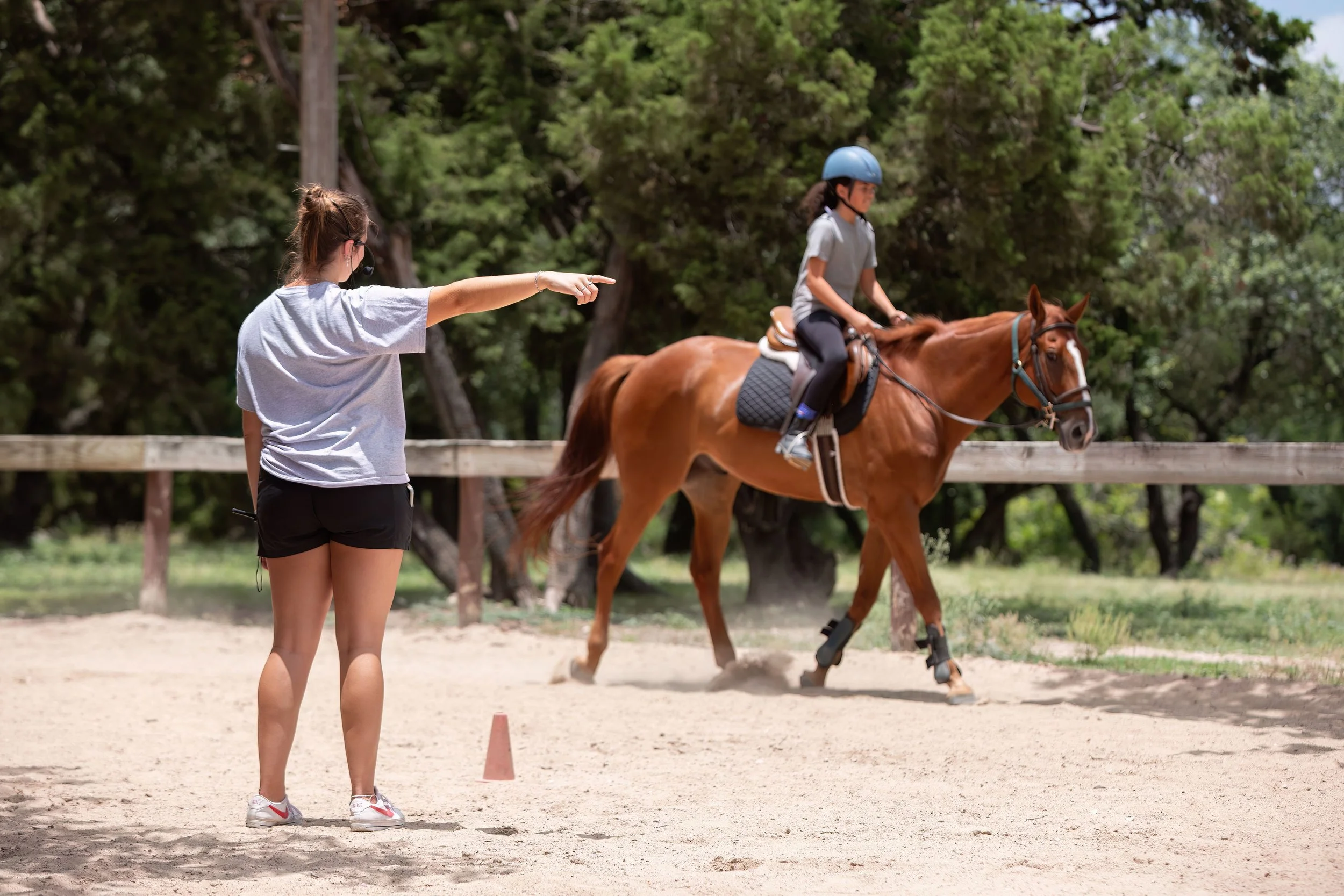 A girl riding a brown horse with a blue helmet during a riding lesson in an outdoor sandy arena. A woman with brown hair tied back, wearing a gray t-shirt, black shorts, and sneakers, points and gives directions.