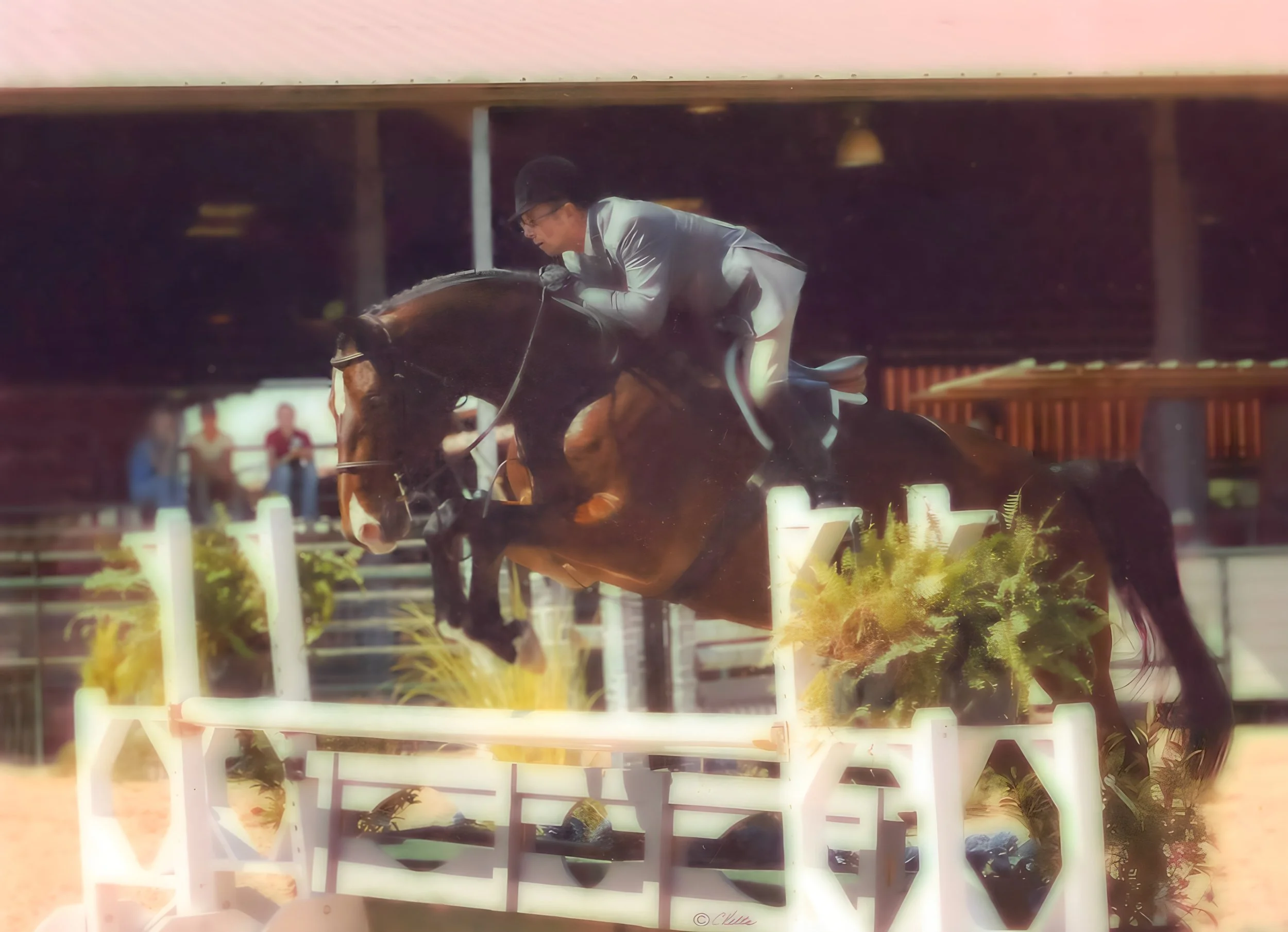 A person wearing a light-colored jacket and dark helmet riding a horse jumping over an obstacle in a show jumping competition.