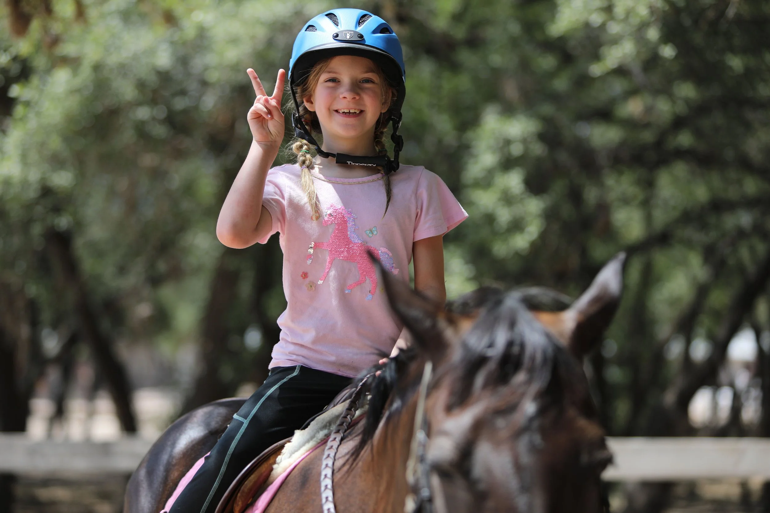 A young girl smiling and making a peace sign while riding a horse outdoors, wearing a blue helmet and a pink t-shirt with a unicorn design.