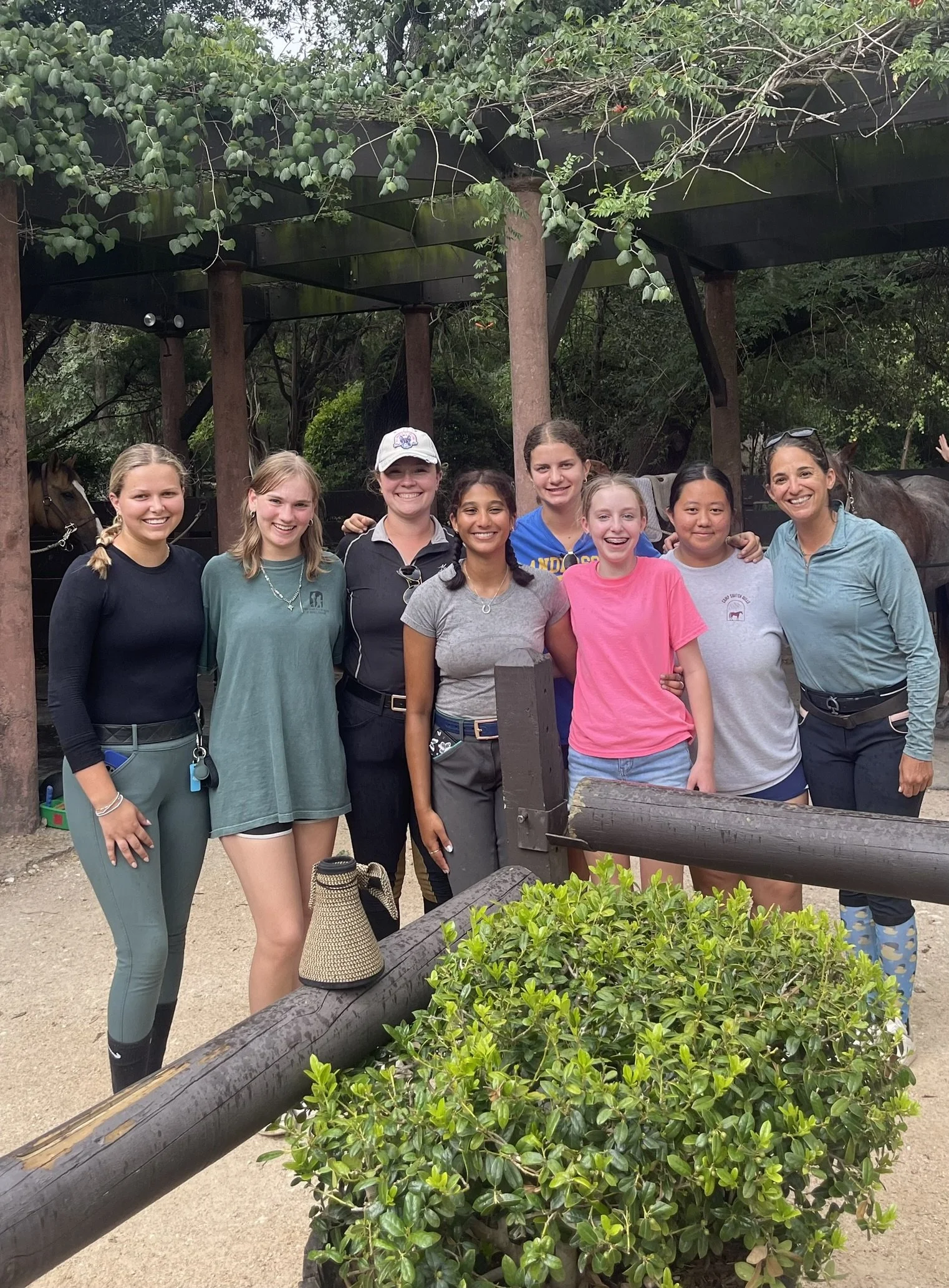 Group of nine women standing together outdoors near some horses, smiling at the camera, trees in the background.