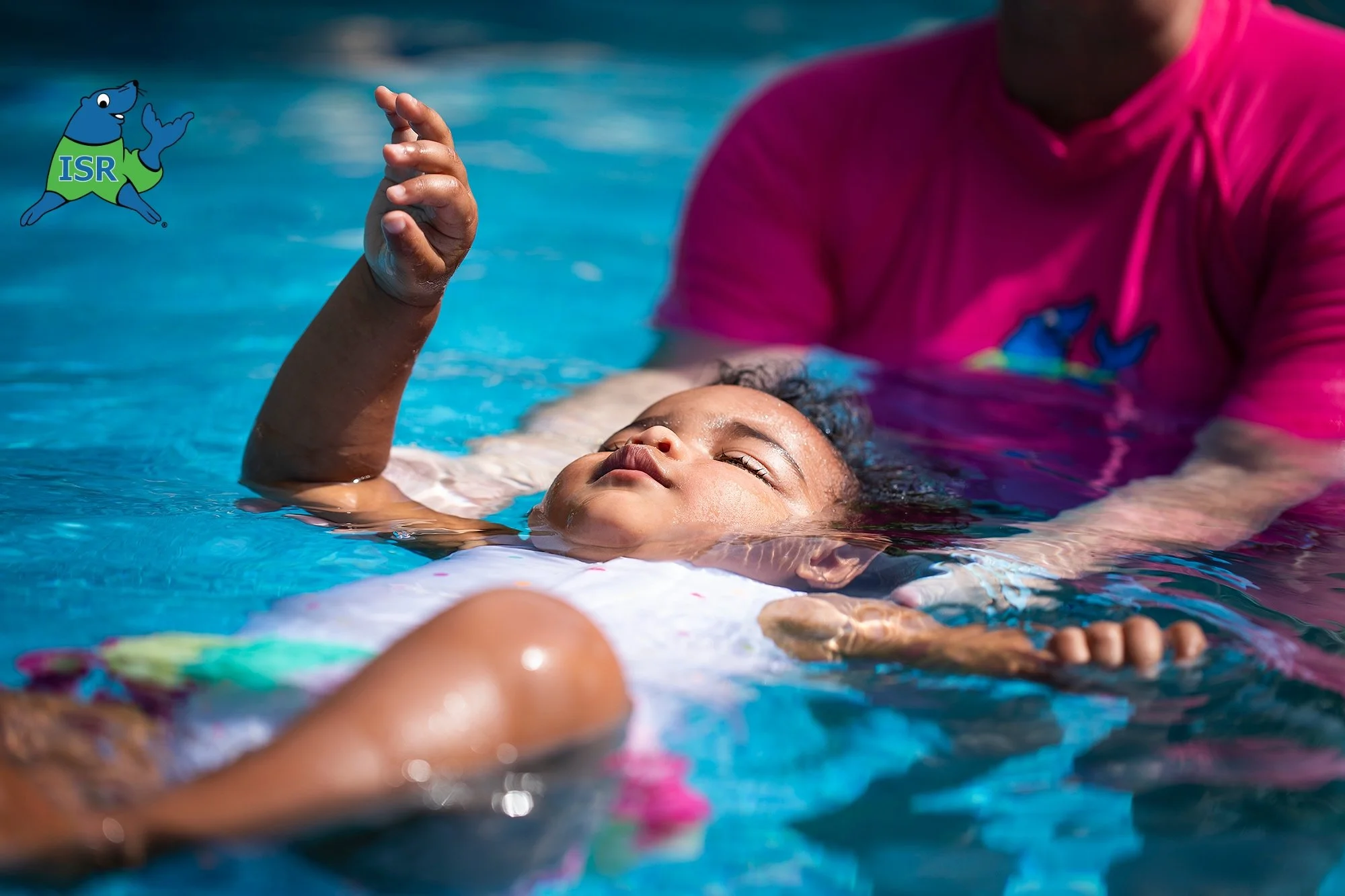A child swimming in a pool with an adult learning to float in ISR survival swim lessons, with an ISR logo on the child's clothing and the adult wearing a pink shirt.