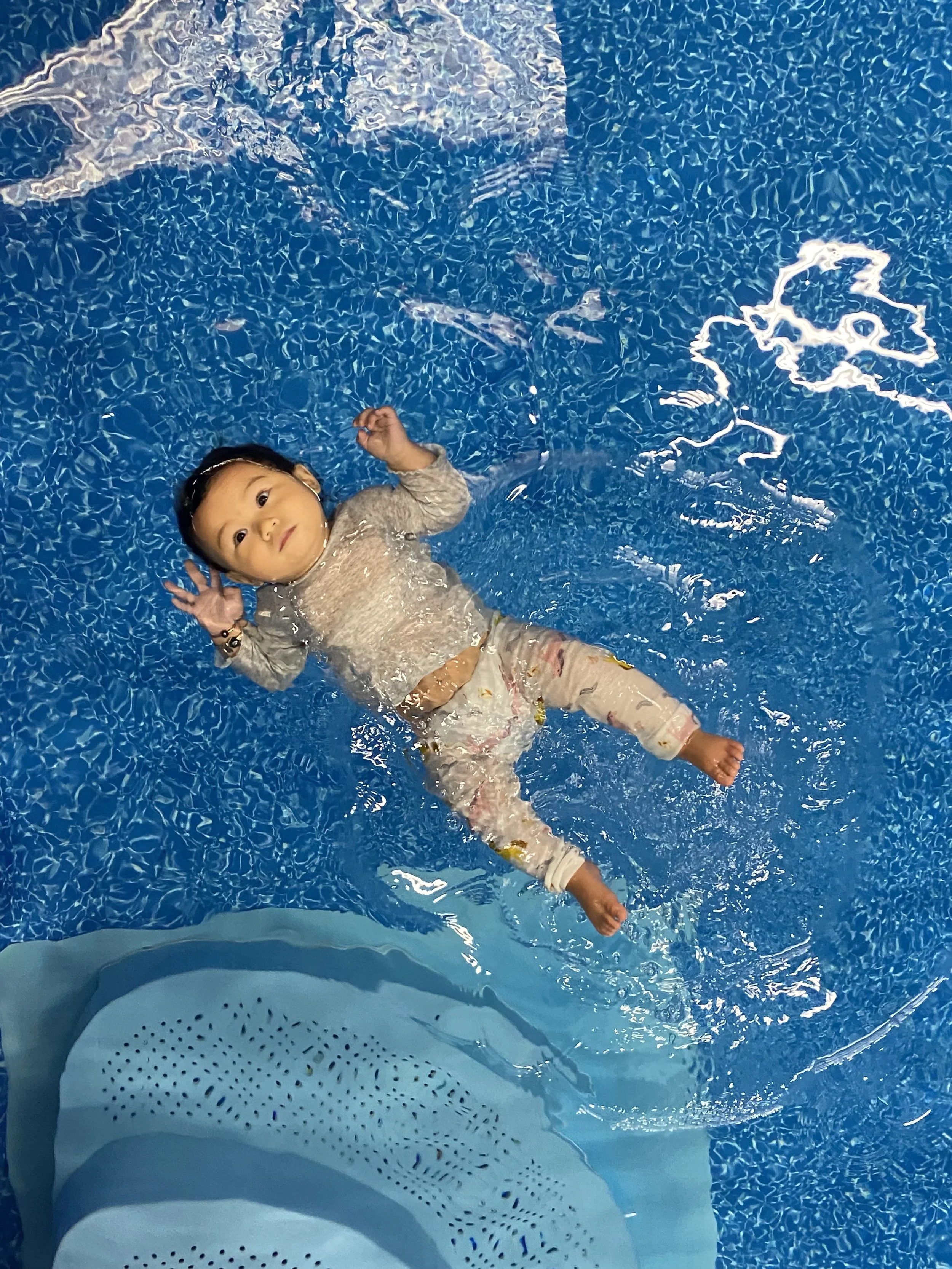 A young girl floating on her back in a swimming pool, wearing a long-sleeve shirt and patterned pants, looking at the camera while holding her hand near her ear. An child showing off her float after completing her ISR Survival Swim lessons.