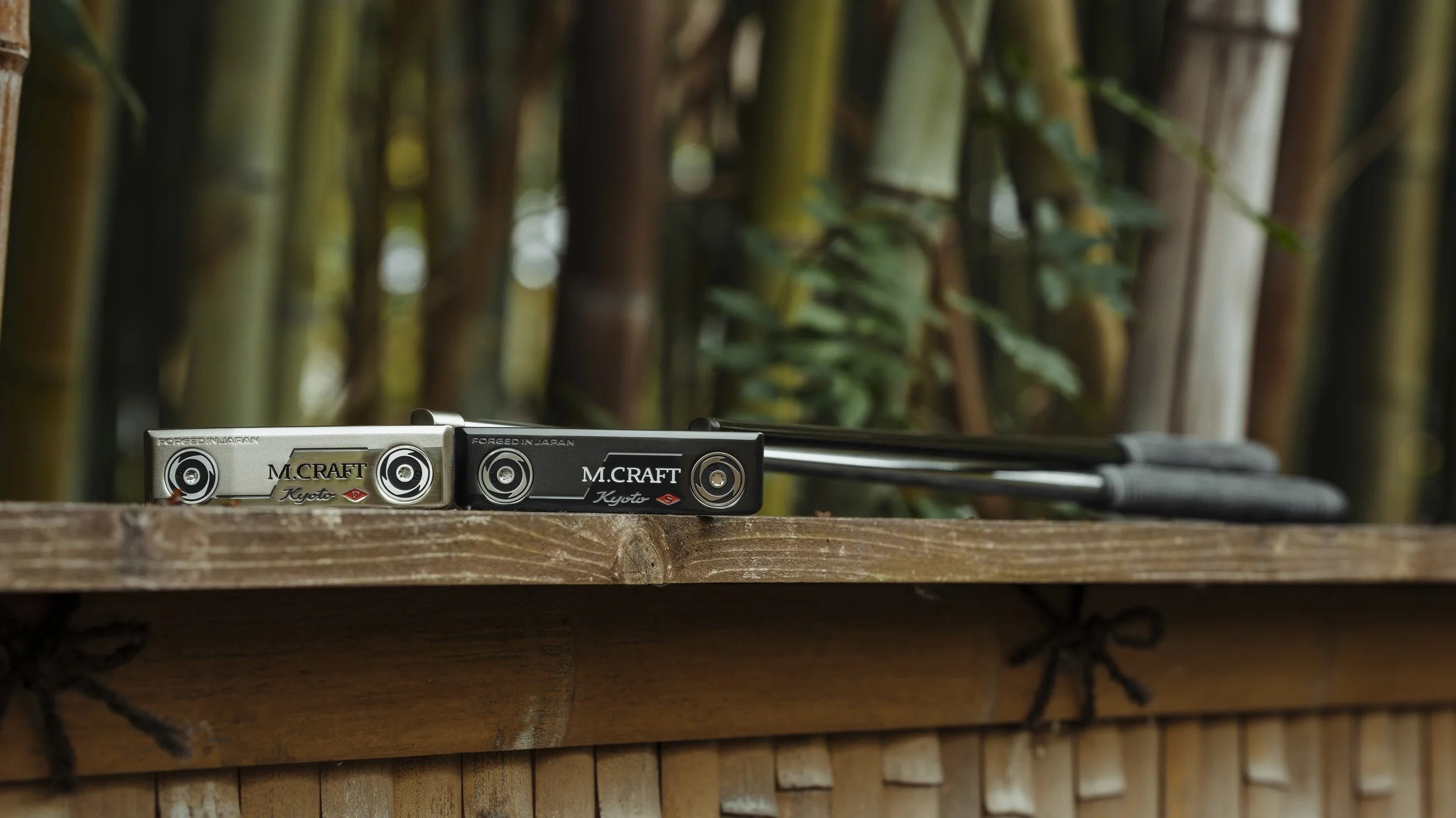 Two golf putters, one silver and one black, resting on a wooden surface outdoors with bamboo plants in the background.