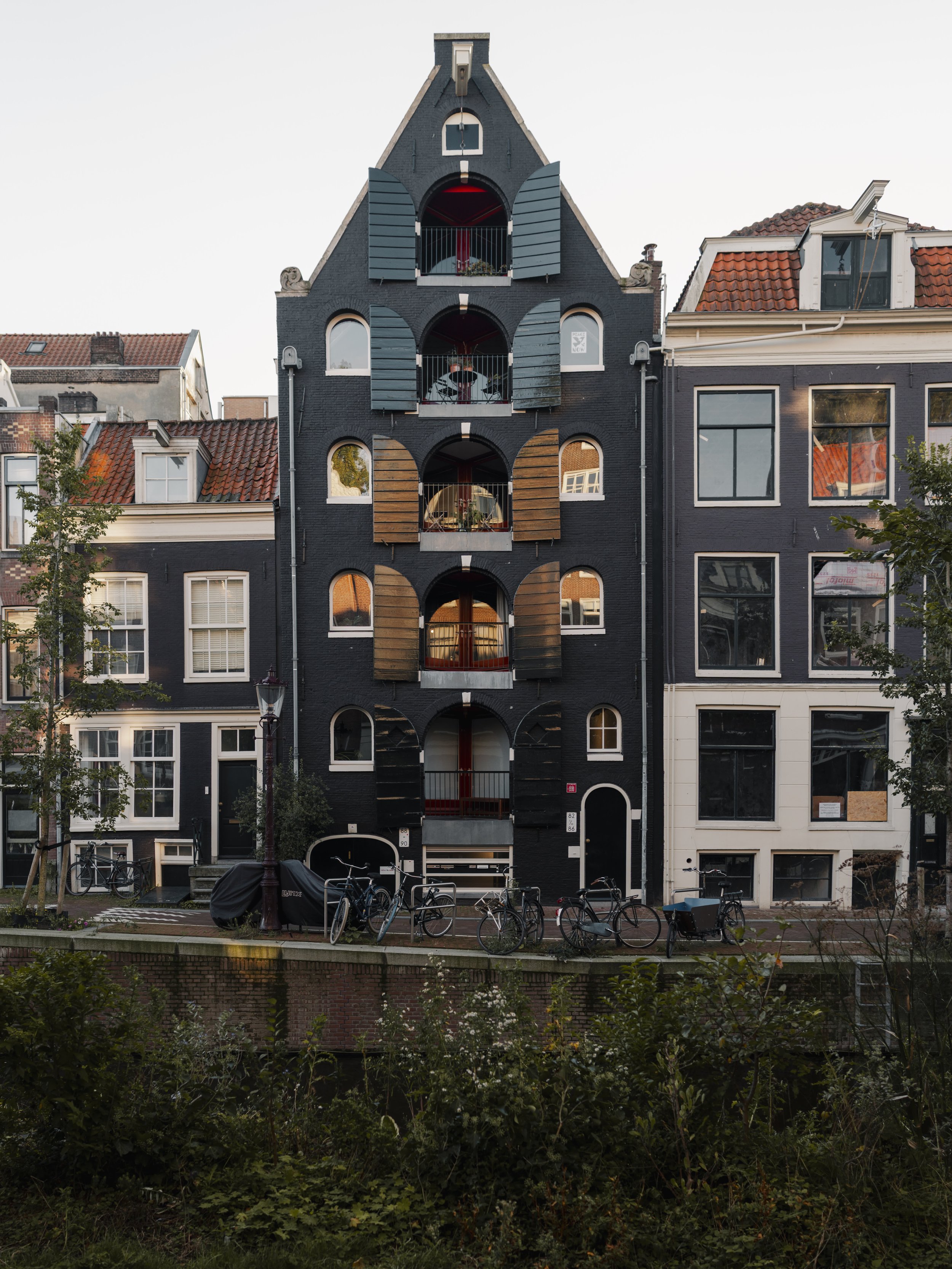 A black multi-story building with unique arched and shuttered windows, located between two other buildings, with several bicycles parked in front on a canal pathway.