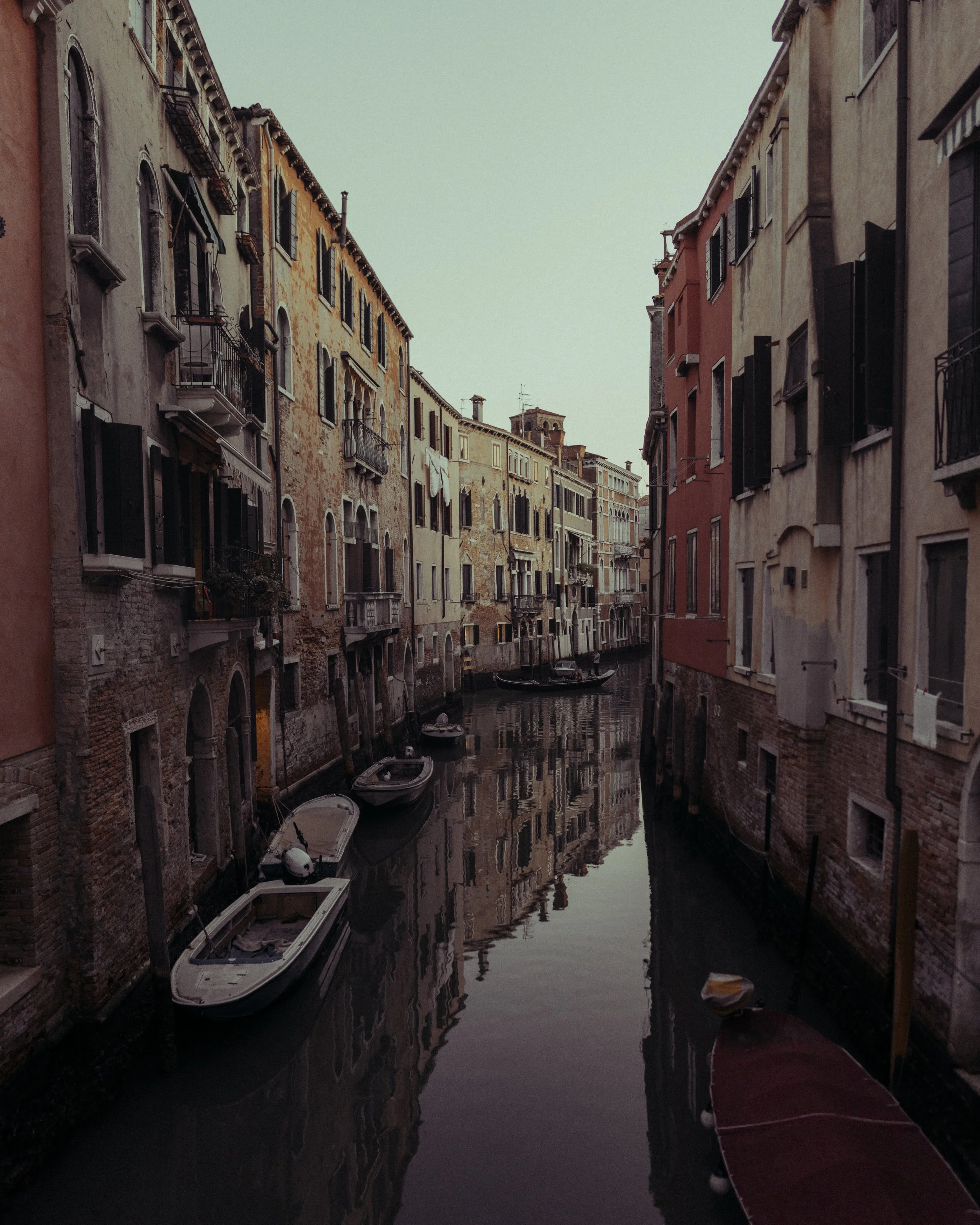 A narrow canal in Venice, Italy, with boats docked along the sides and old colorful buildings with shuttered windows on either side.