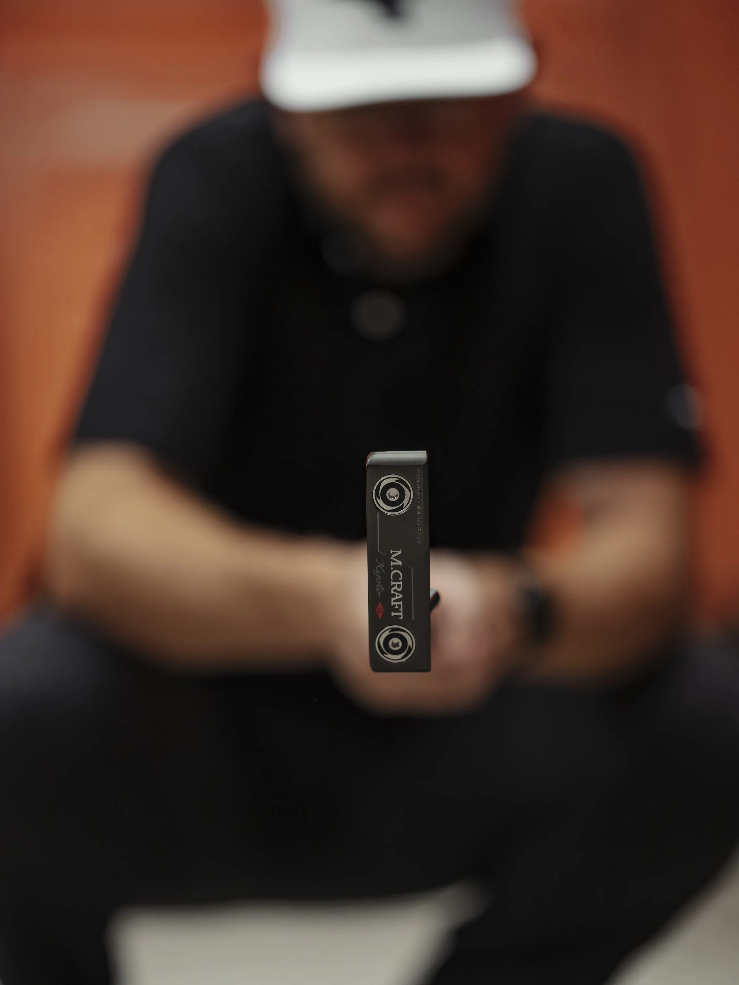 Close-up of a person holding a golf club with the brand 'M.CRAFT' visible on the bottom of the club, focusing on the club face, while the person is blurred in the background.