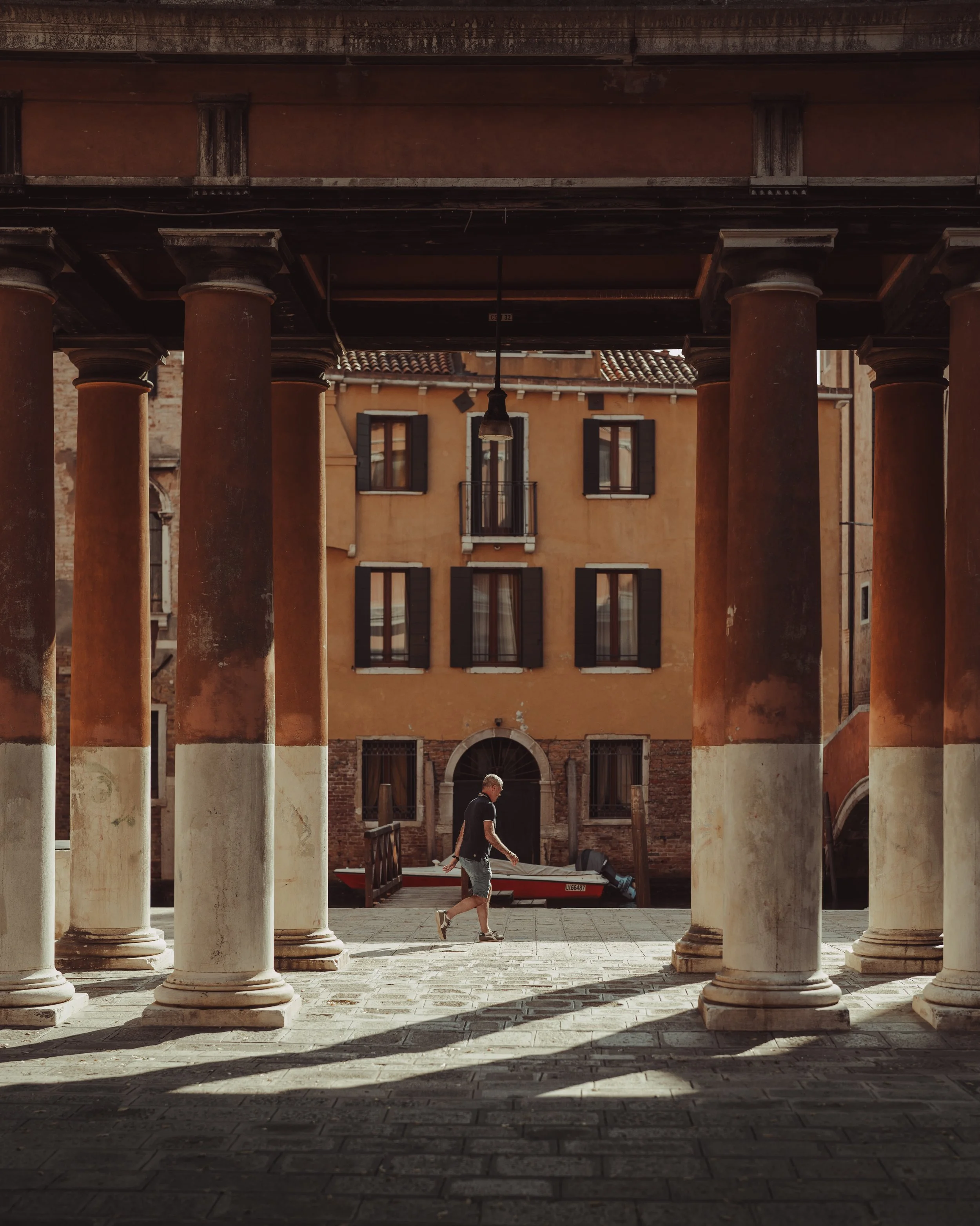 A man walking in an outdoor corridor with large columns, overlooking a canal with a boat and yellow buildings with black shuttered windows in Venice, Italy.