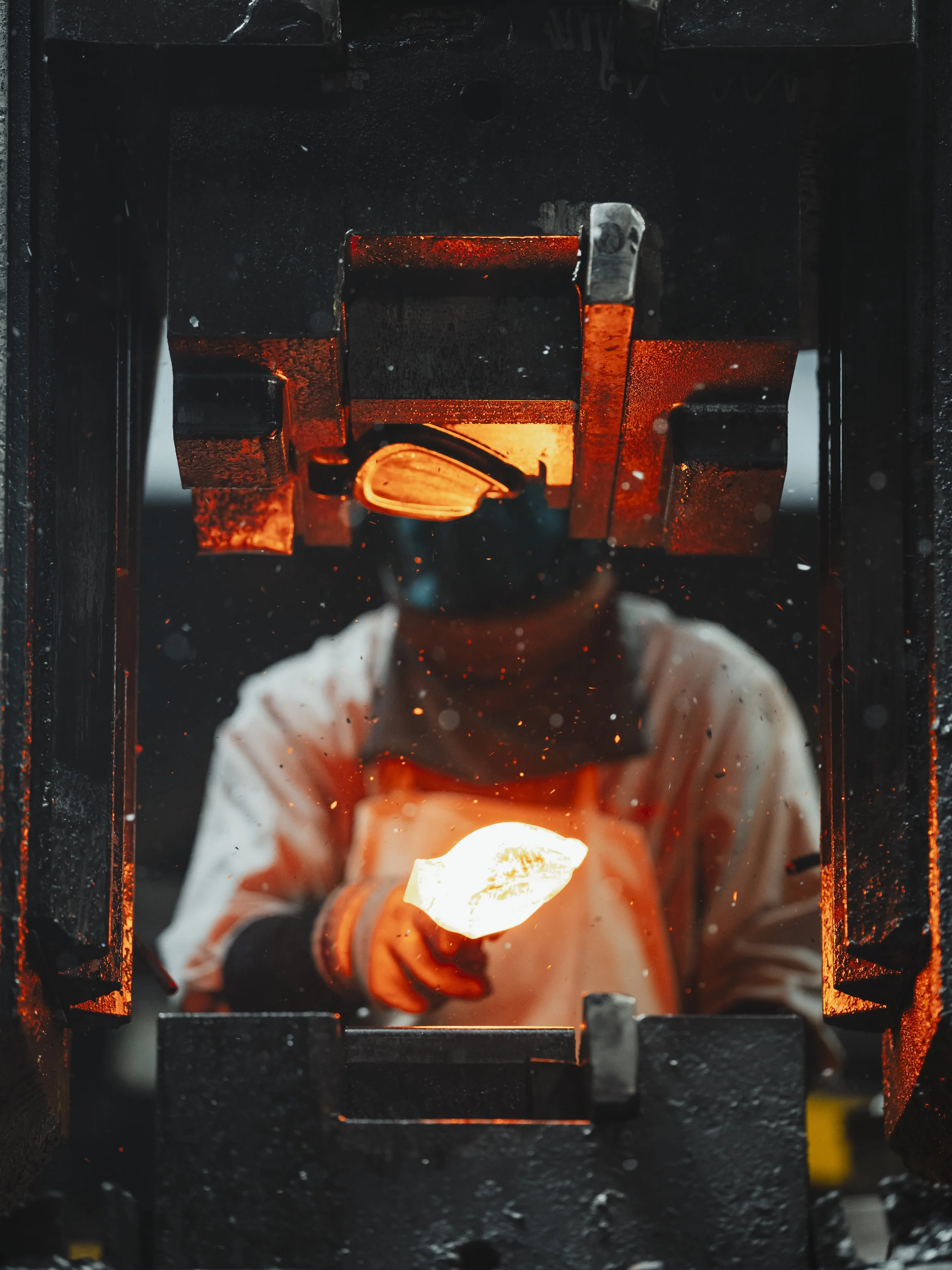 A glassblower wearing protective gear, including a face shield and gloves, working with molten glass in a furnace.